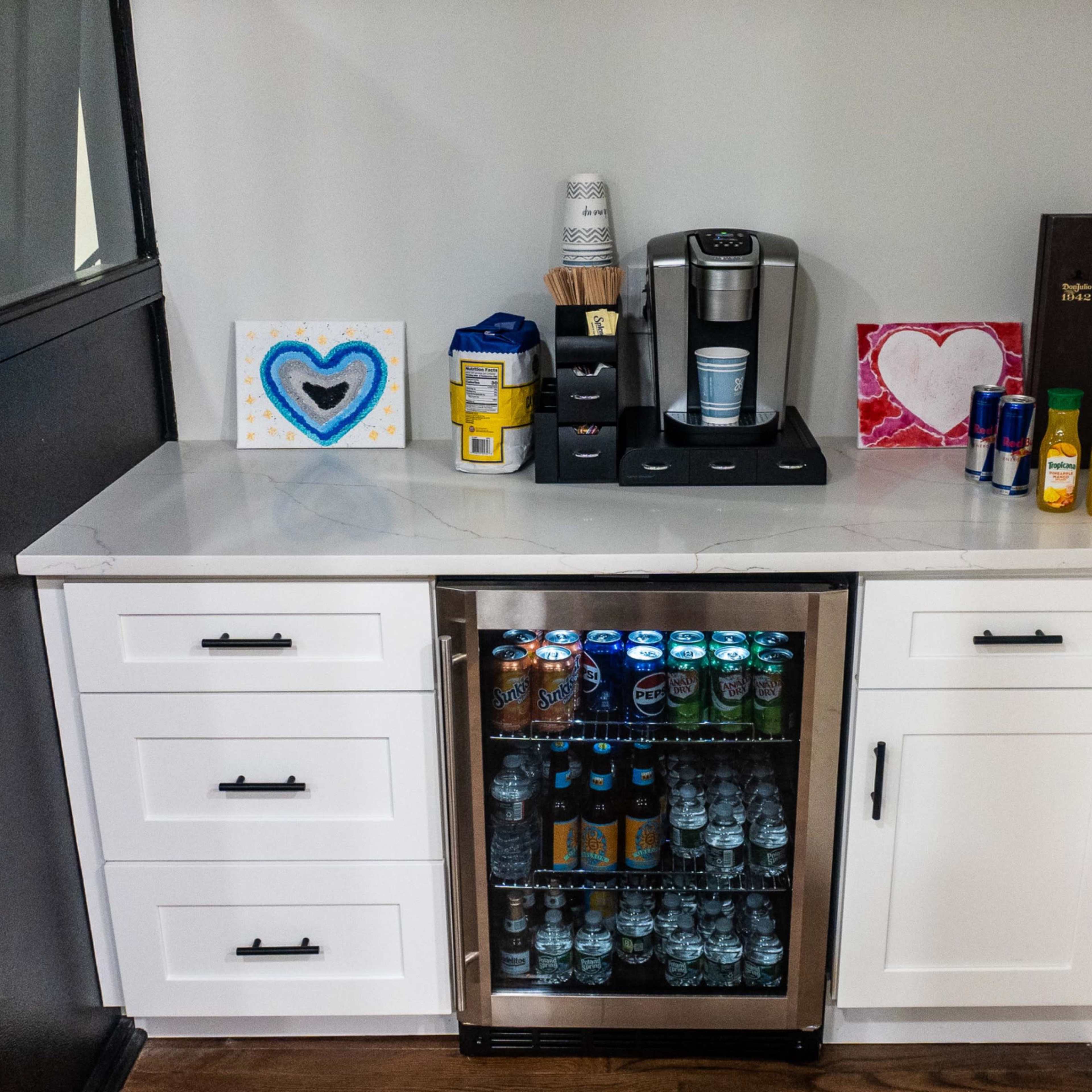 A small kitchen area with a countertop, under-counter refrigerator stocked with beverages, and various items including a coffee maker, cups, and decorations.