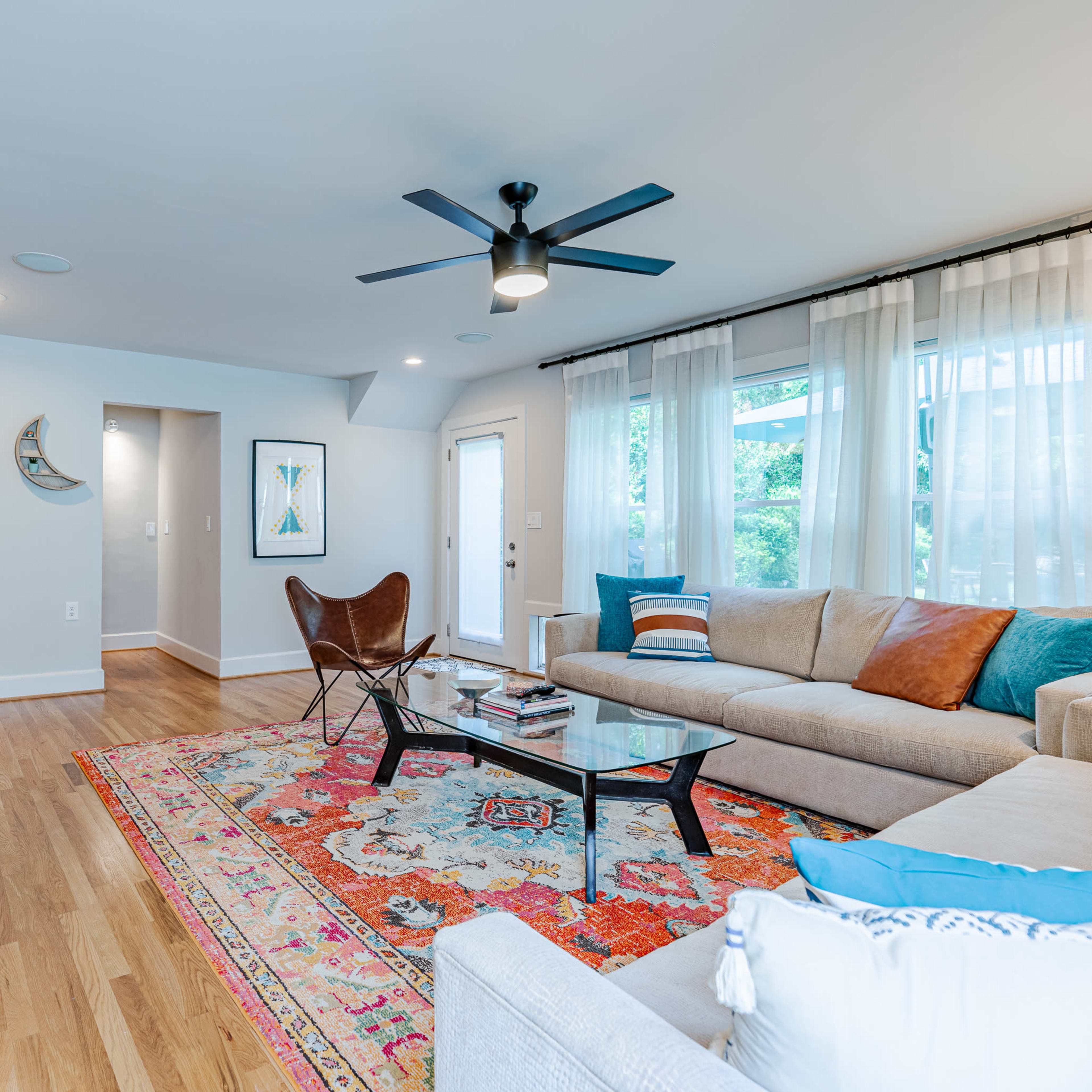 A modern living room features a sectional sofa, a butterfly chair, a glass coffee table, and a colorful area rug, with large windows allowing natural light to enter.