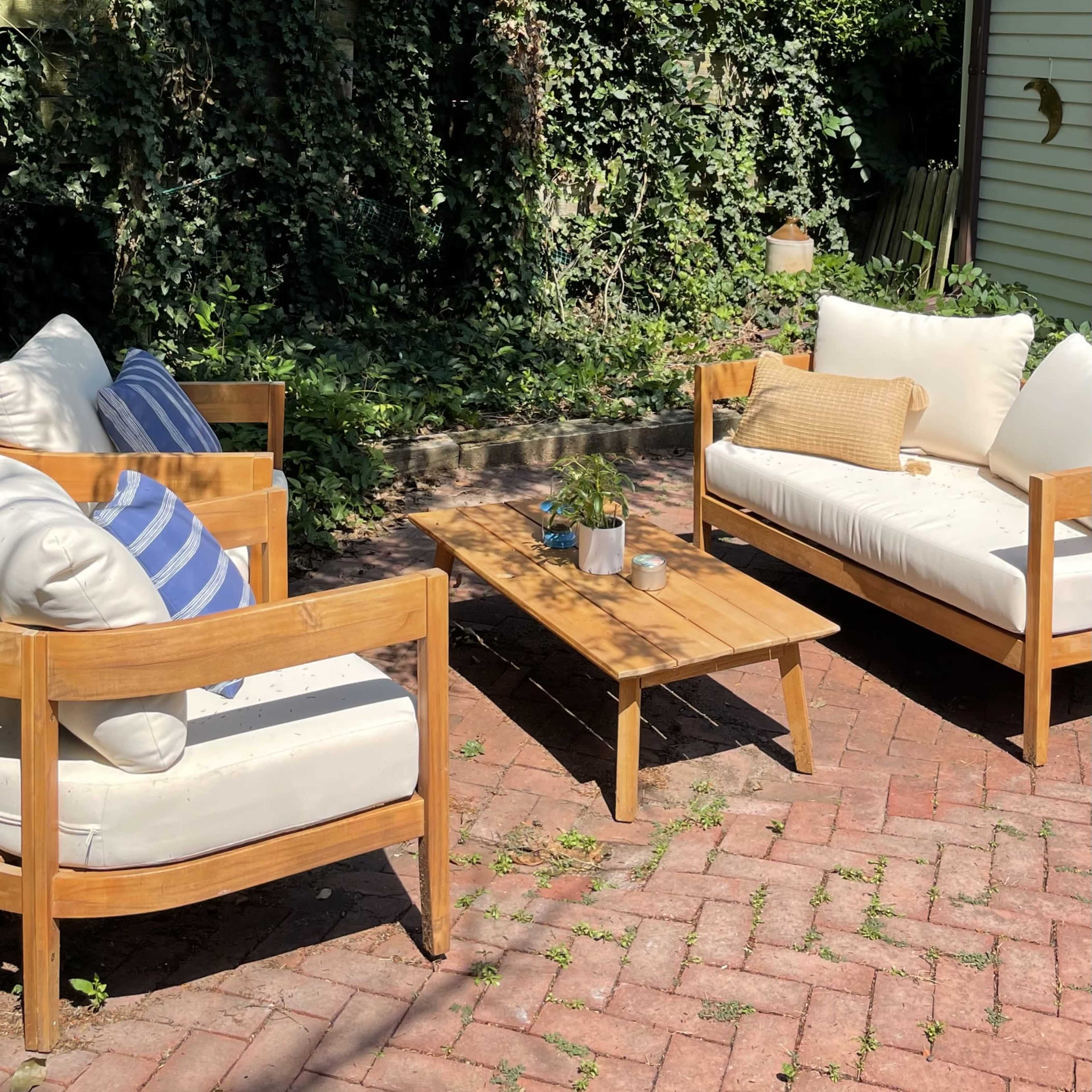 A small patio area features two armchairs and a two-seat sofa arranged around a wooden coffee table amidst a green backdrop of plants and trees.