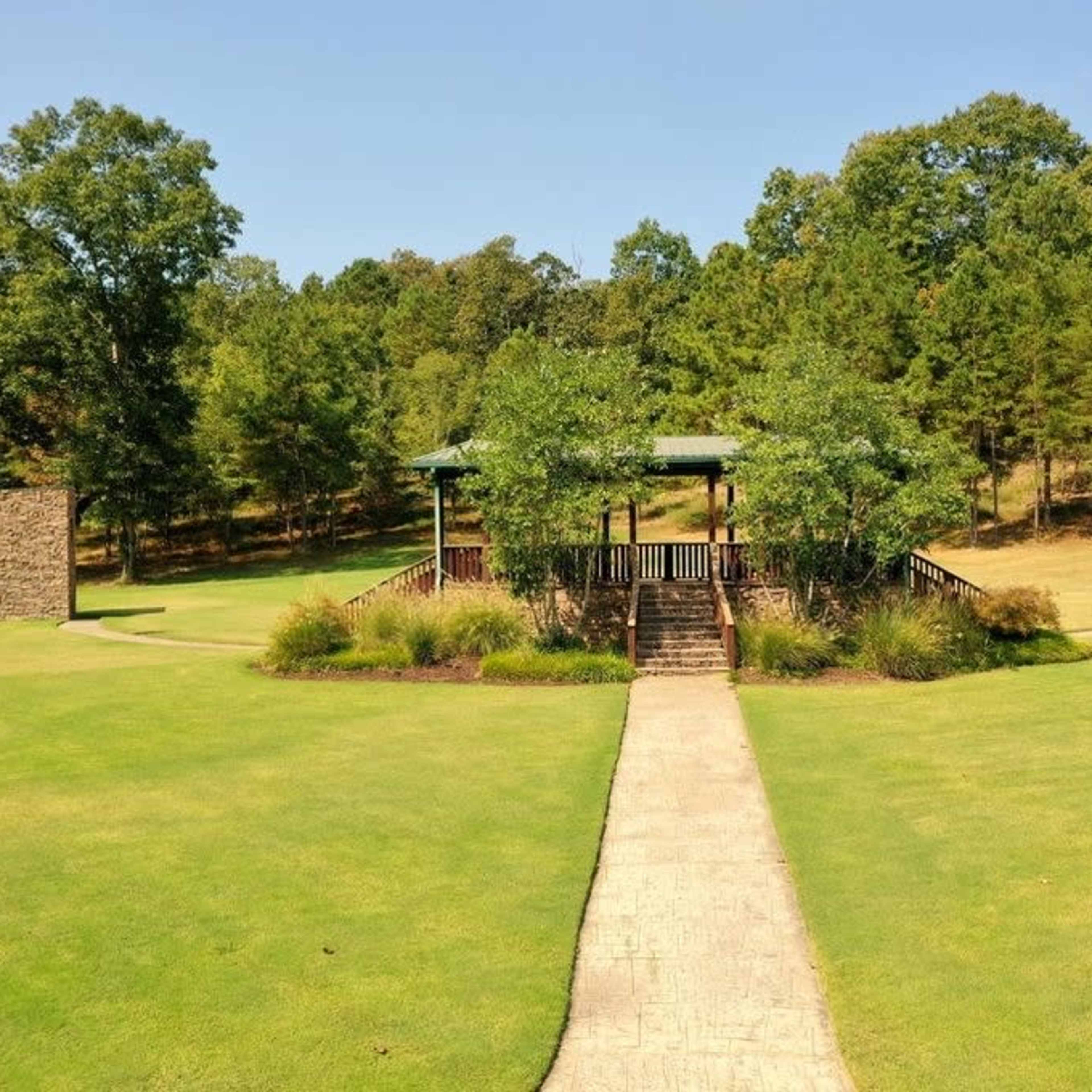 A concrete pathway leads to a covered gazebo surrounded by landscaped greenery in a park setting.