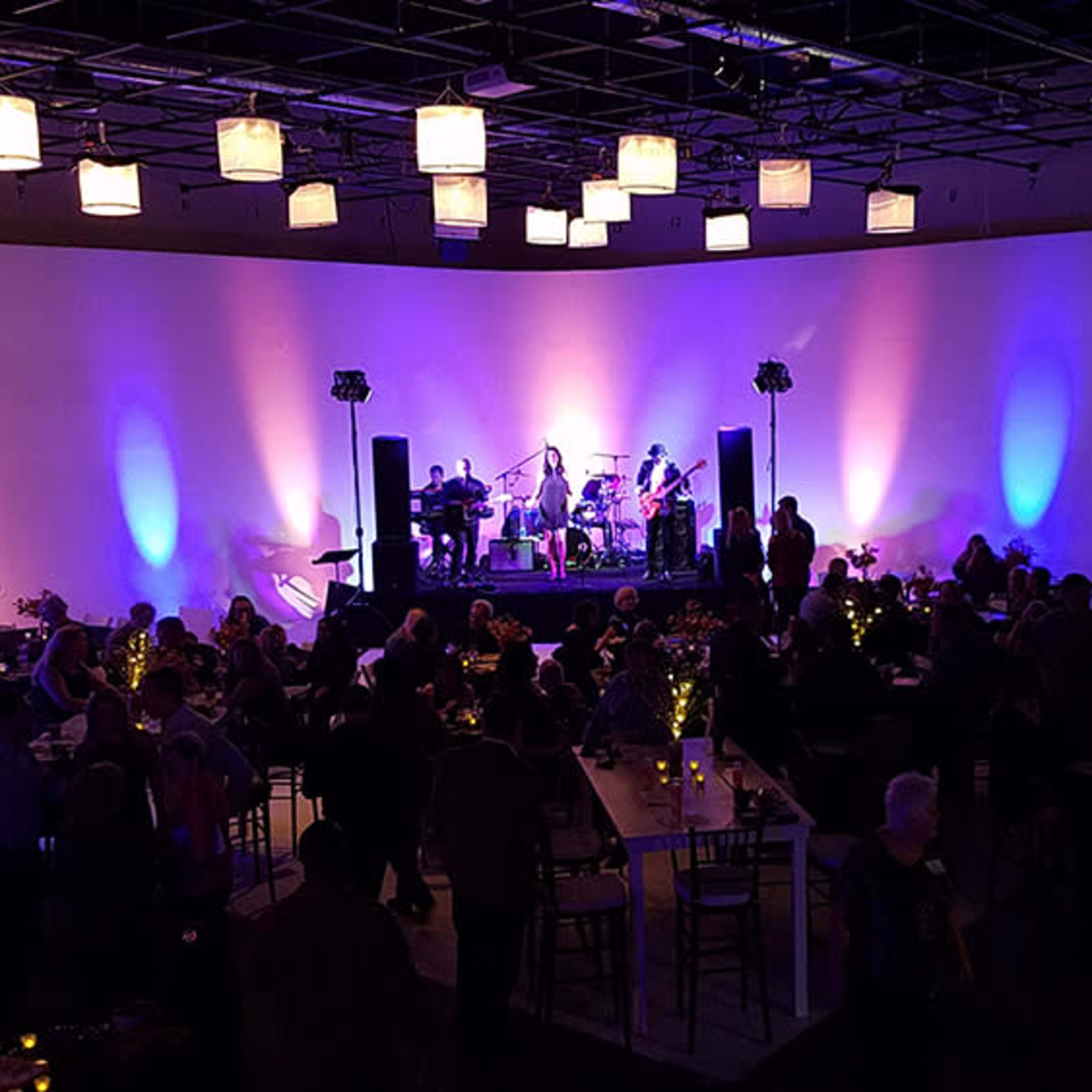 A large crowd gathers indoors for an event featuring a band on stage, with soft lighting and decorative tables.