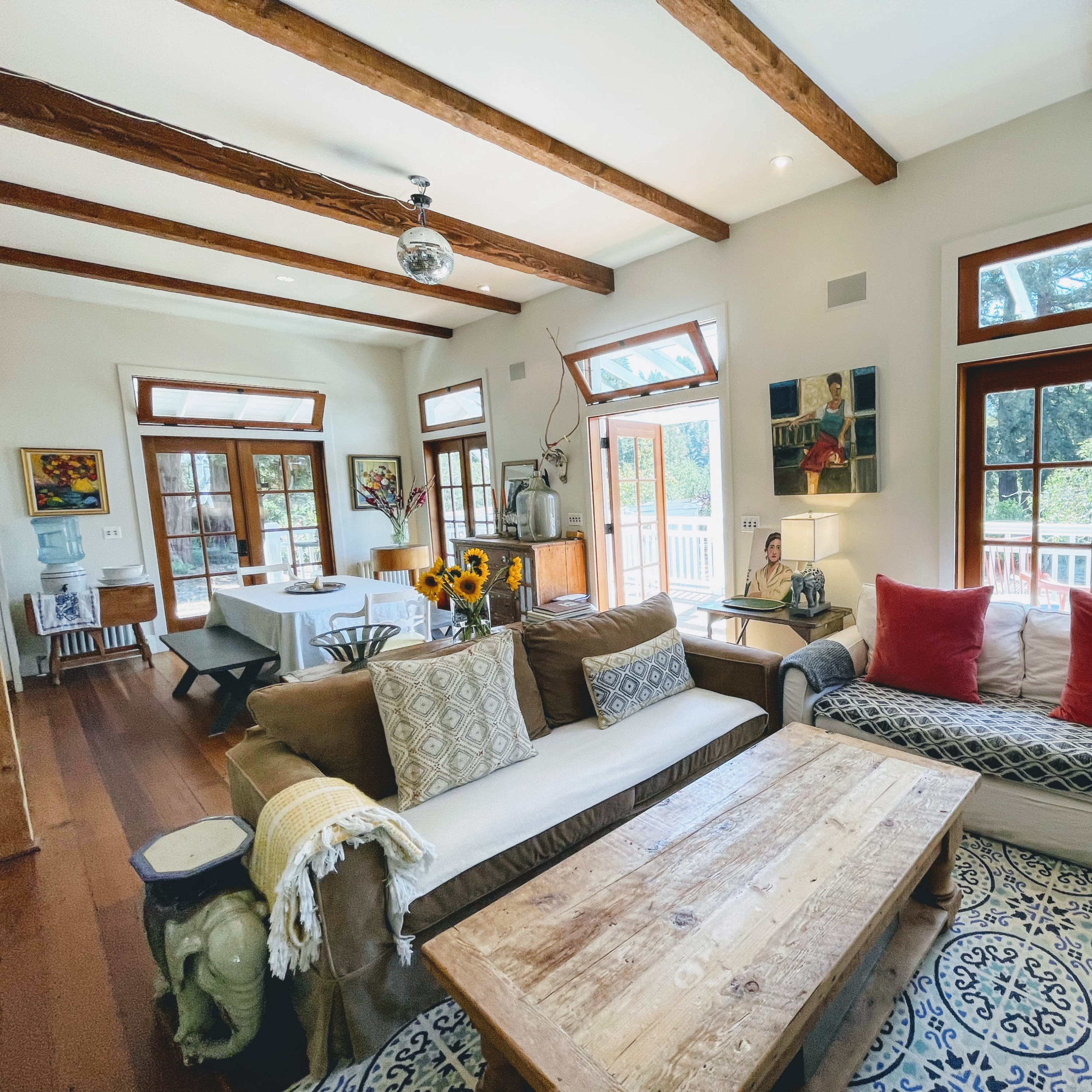 A bright living room features wooden beams, large windows, a rustic wooden coffee table, and various pieces of furniture arranged around a patterned rug.