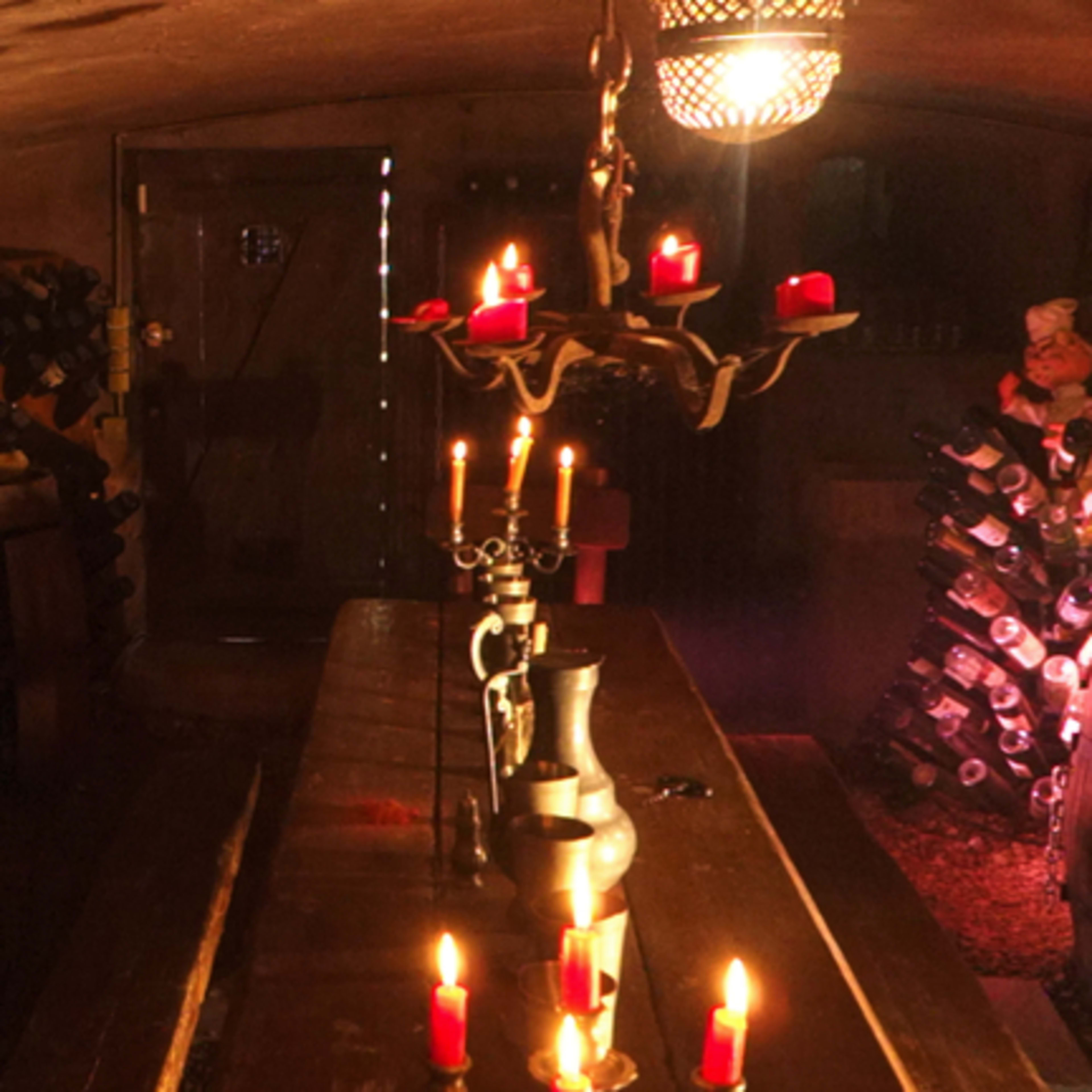 A dimly lit wine cellar with wooden shelves filled with bottles, a table at the center, and a chandelier with candles providing illumination.