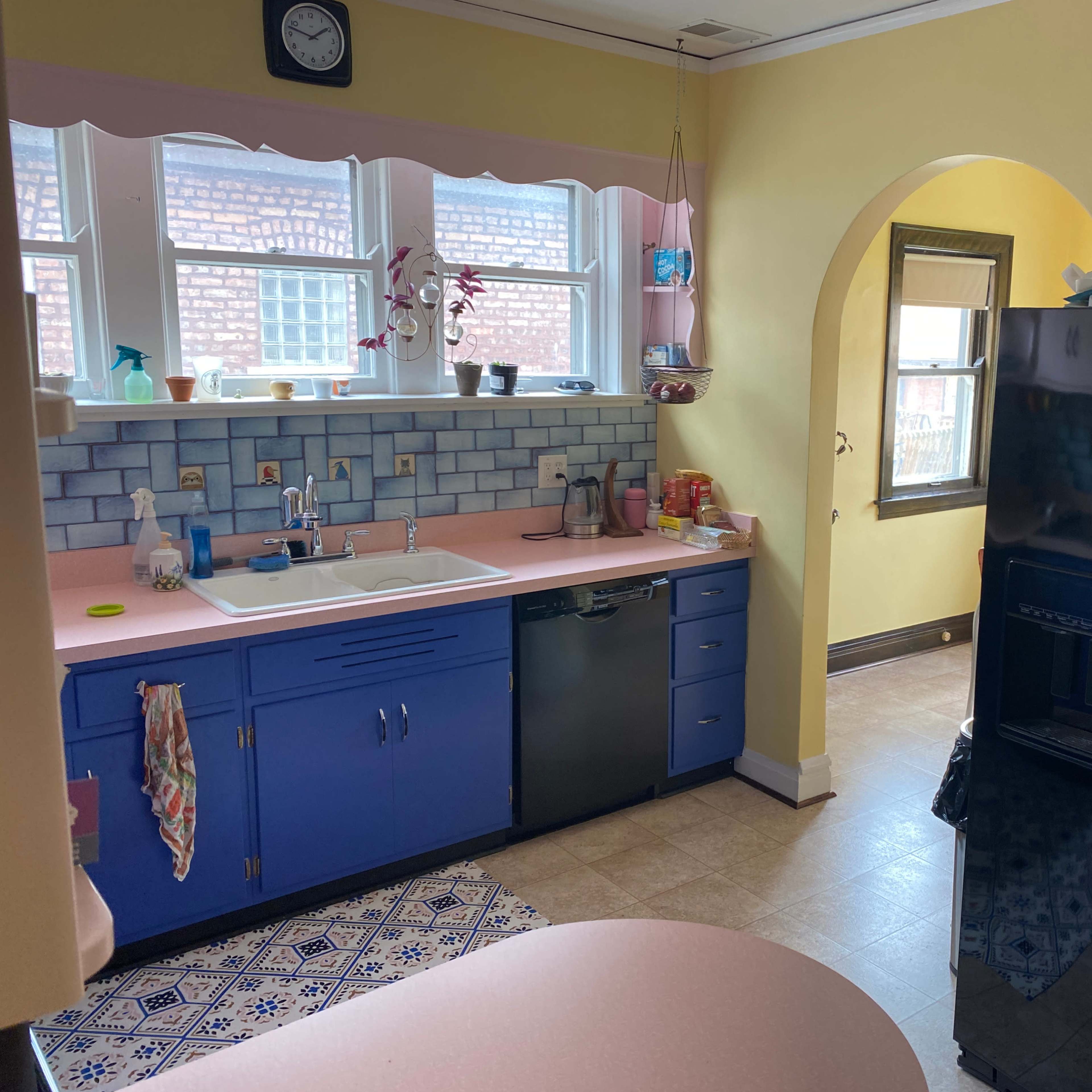 A kitchen with blue cabinets, a black refrigerator, a pink countertop, and a window that lets in natural light.