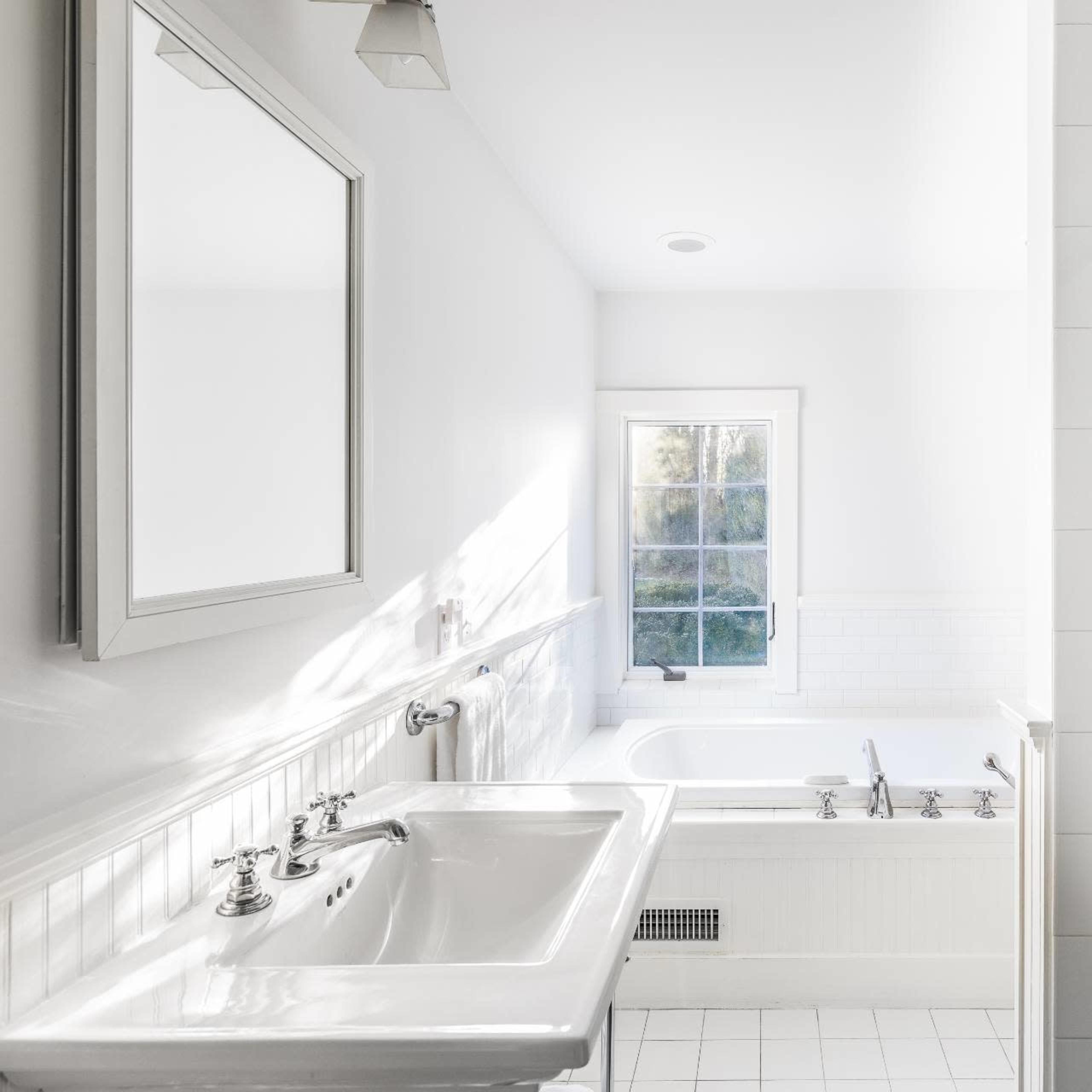 A bright bathroom features a sink and large mirror on one side, a shower stall on the other, and a window overlooking a serene outdoor view.