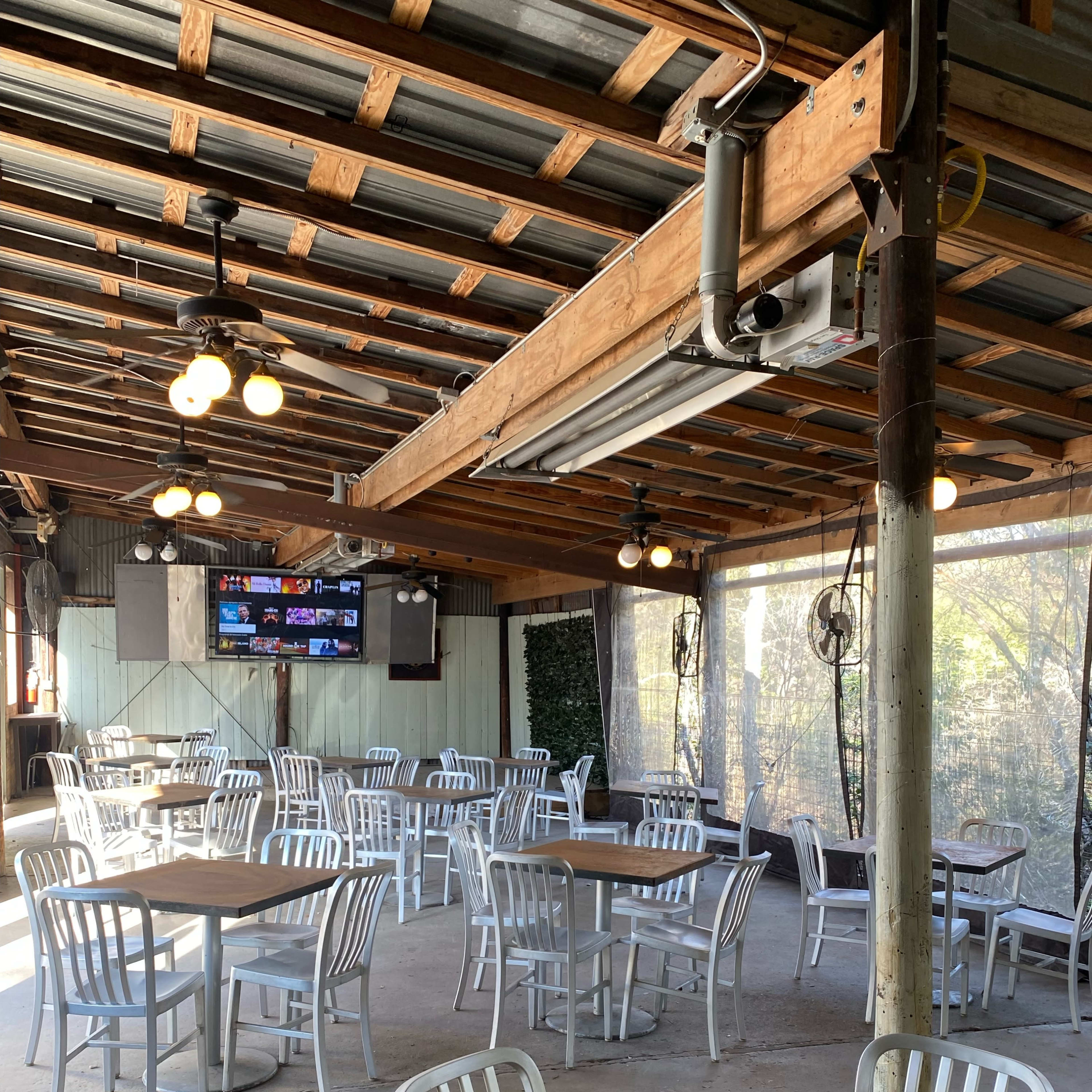 An empty dining area features several white tables and chairs beneath a metal roof with exposed beams and soft lighting.
