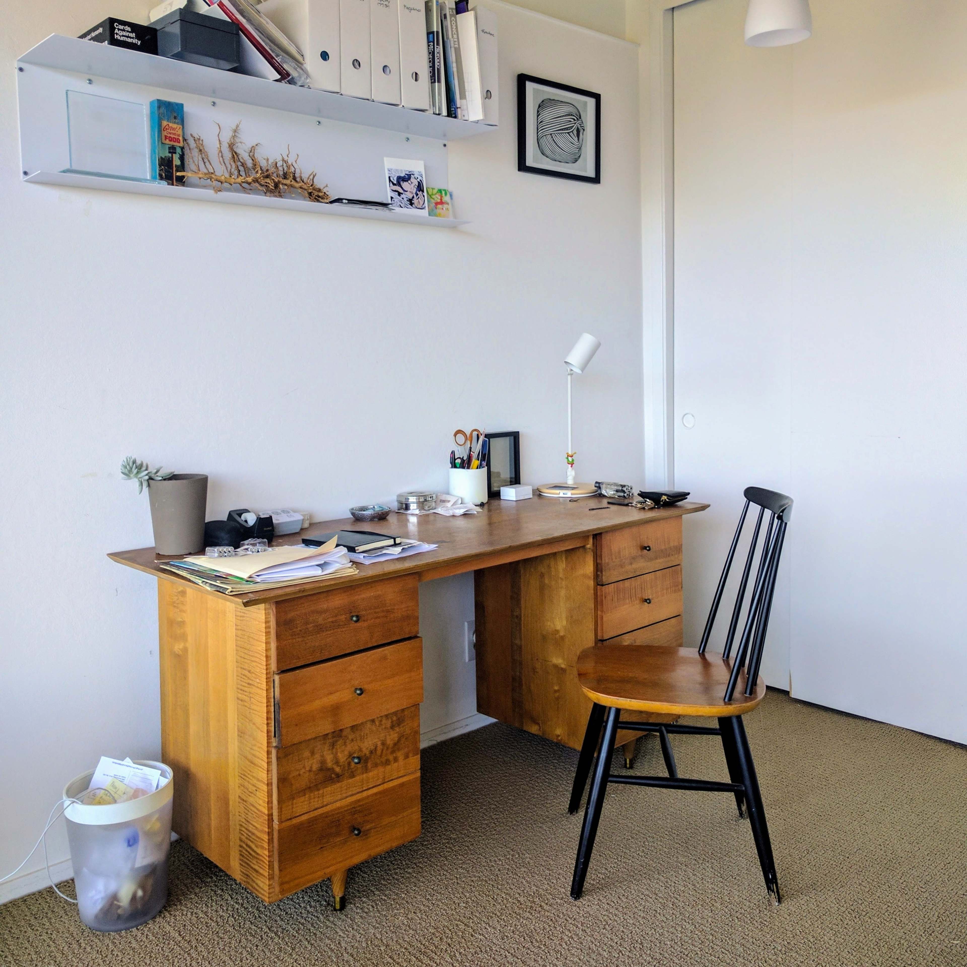 A wooden desk with a chair sits against a wall, accompanied by shelves holding various items and a closed closet door nearby.
