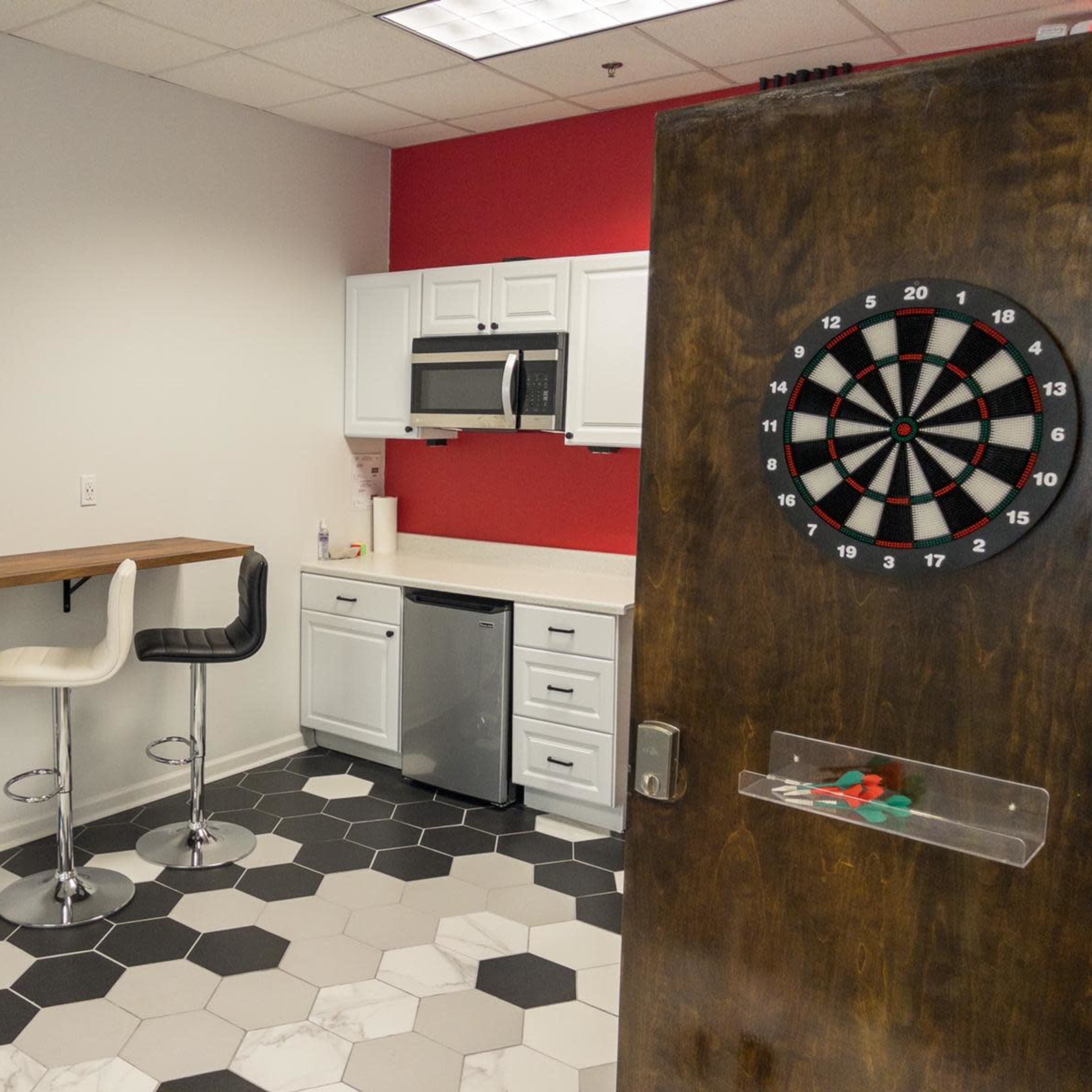 A kitchenette area features two bar stools, a microwave, and a dartboard on a wooden door against a red wall.