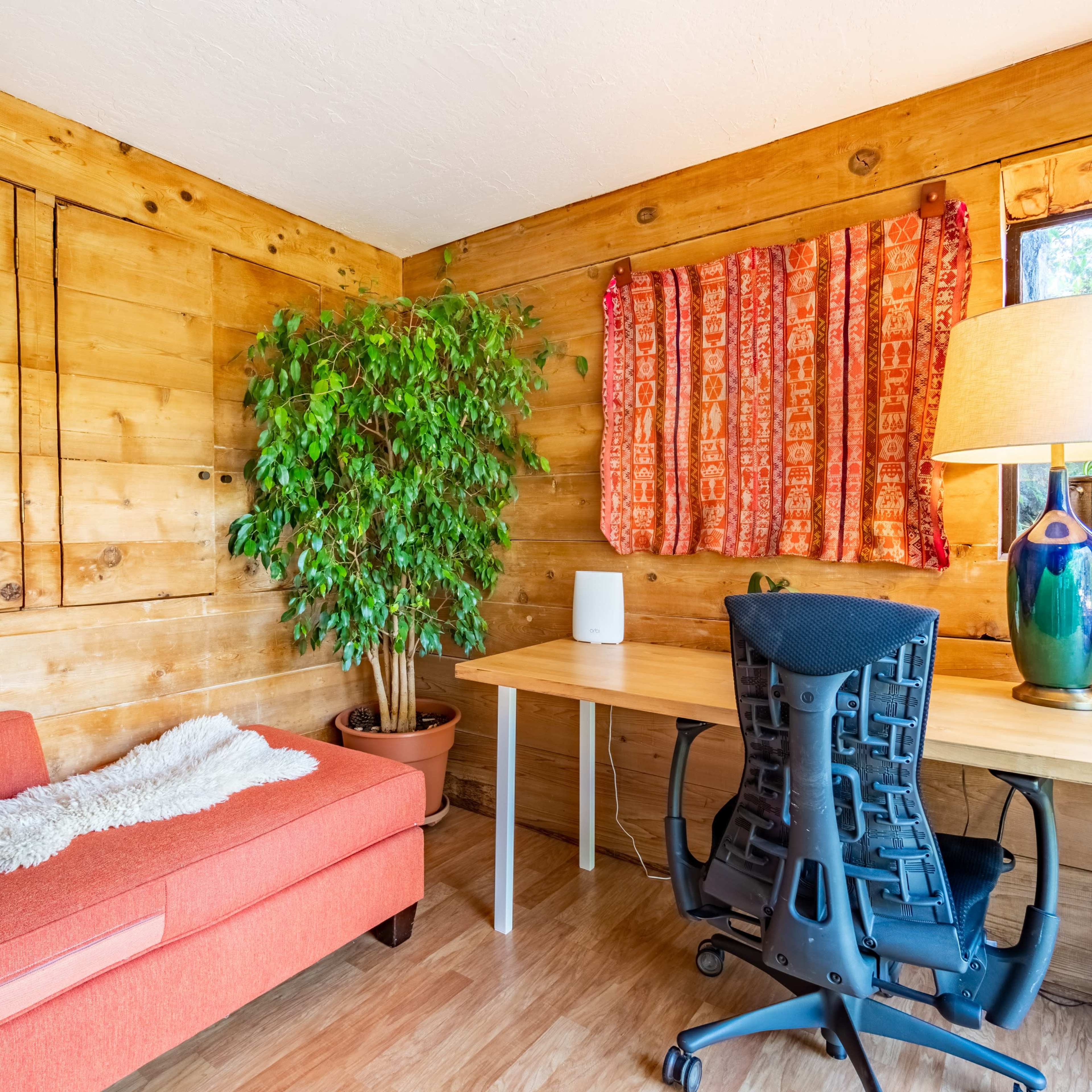 A small wooden room featuring a green plant, a desk with a chair, and a red couch with a decorative throw.