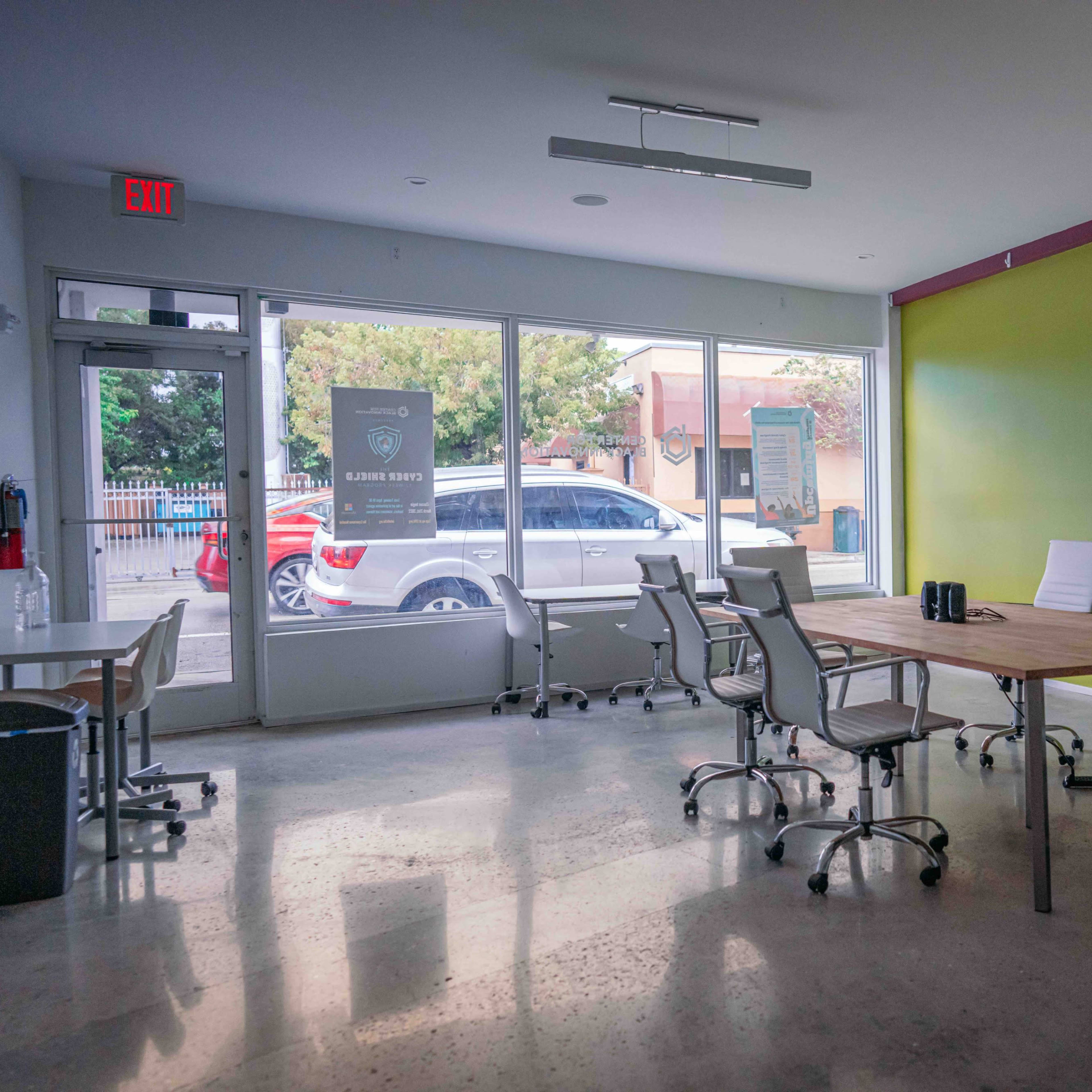 An empty meeting room with a large wooden table surrounded by white chairs, set against a green accent wall and large windows.