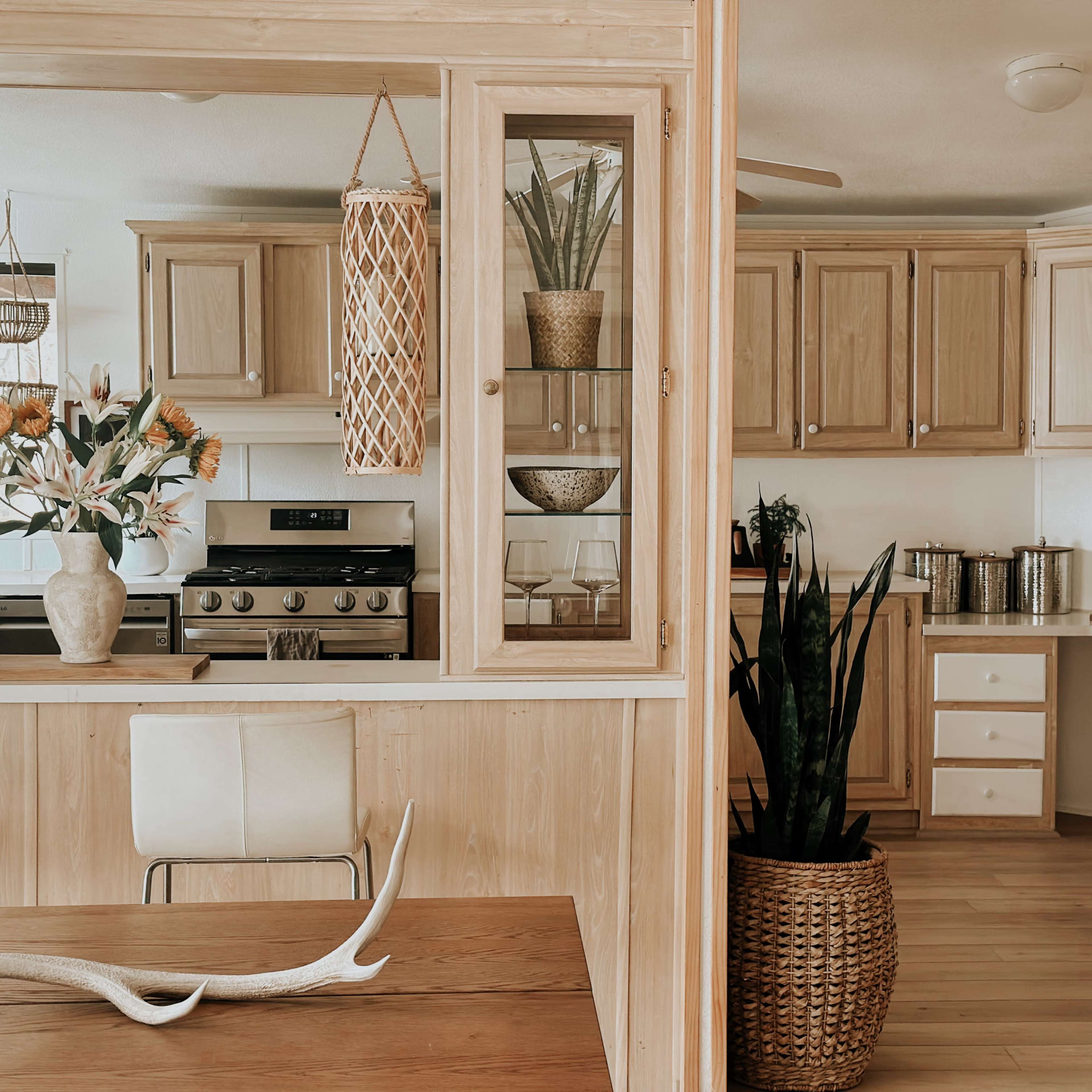 A light wood-furnished kitchen and dining area, featuring a gas stove, potted plants, and a dining table with decorative elements.