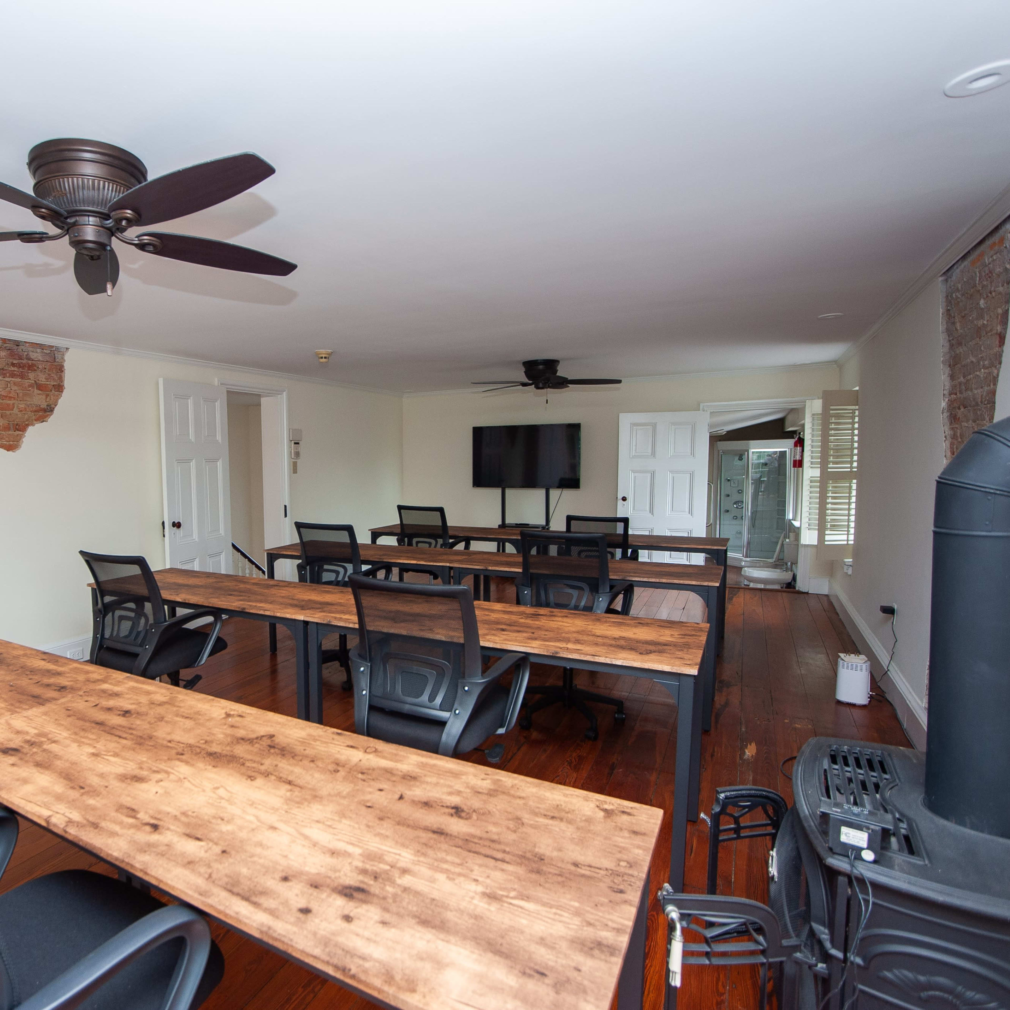 A meeting room with several wooden tables and chairs, a ceiling fan, and a television mounted on the wall.