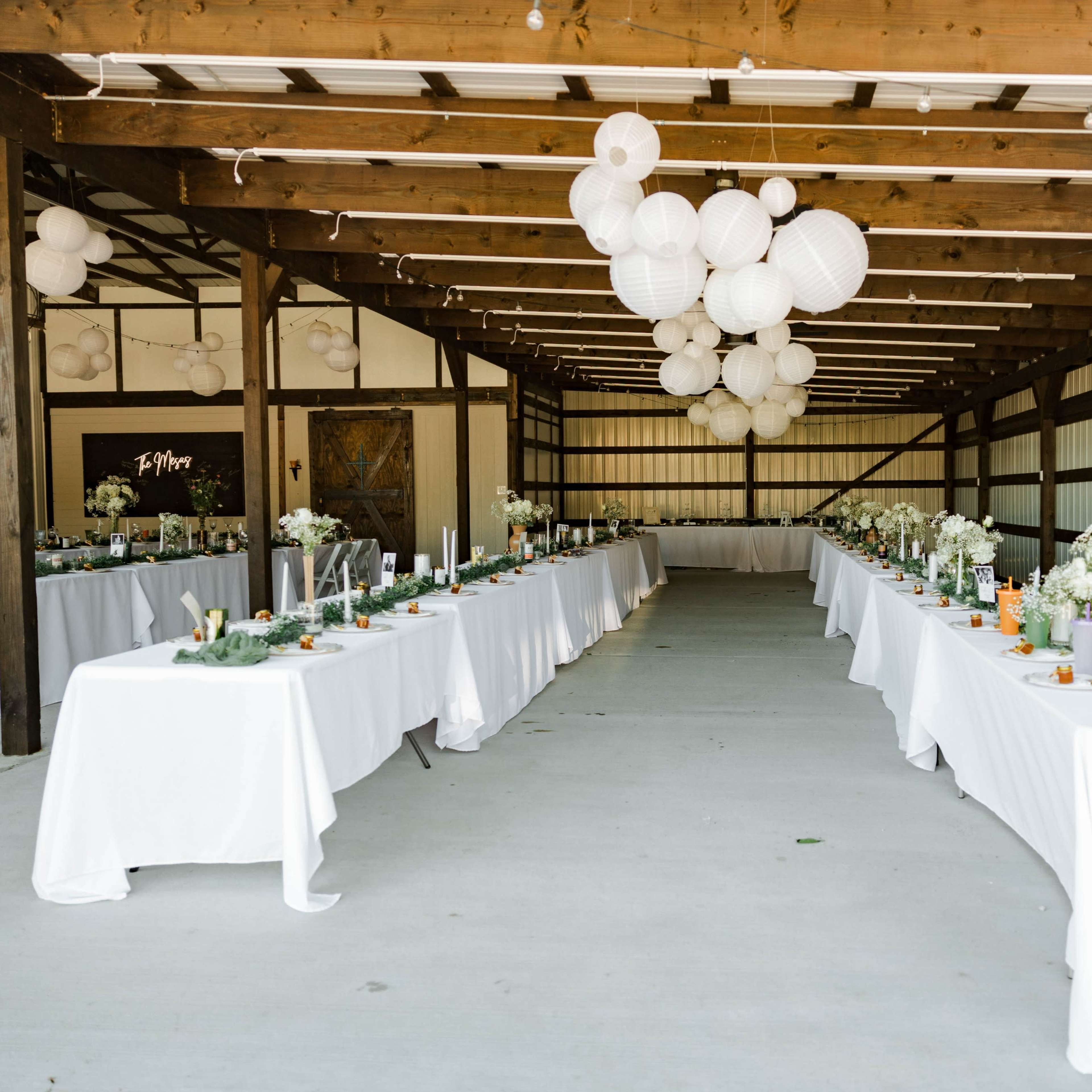 A spacious barn setting features two long tables adorned with white tablecloths and floral centerpieces, illuminated by decorative paper lanterns overhead.