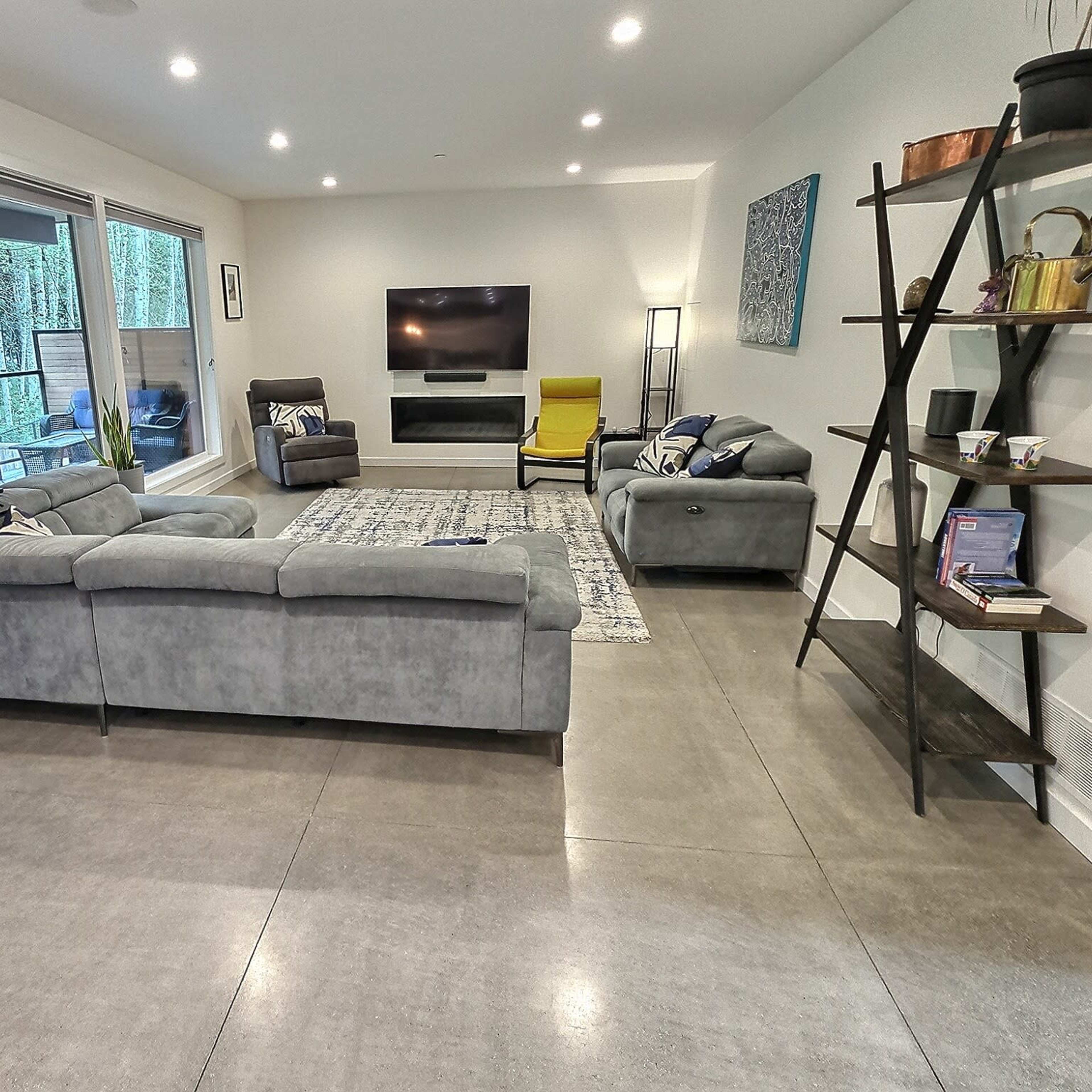 A modern living room with gray furniture, a television mounted on the wall, and large windows facing an outdoor area.
