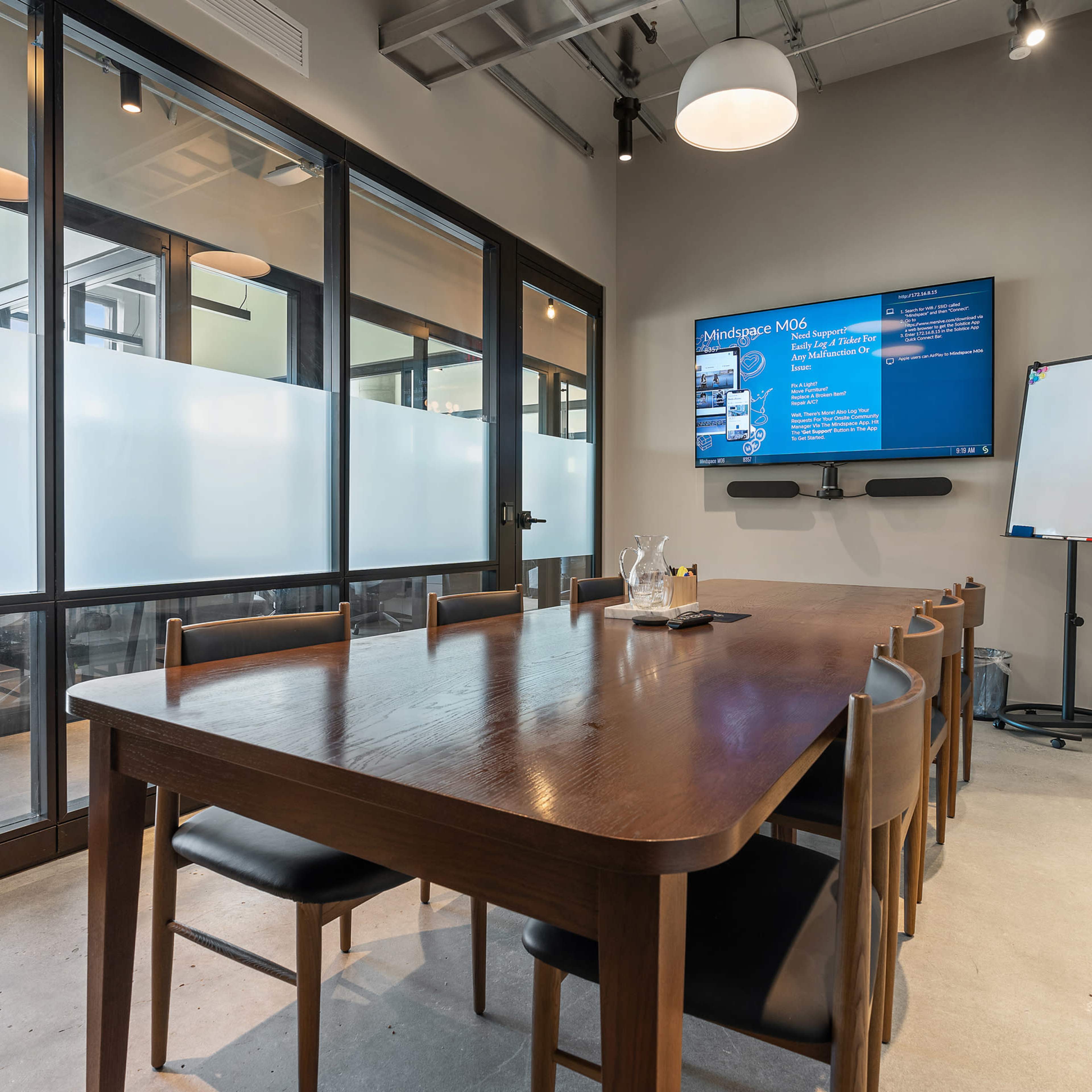 A modern conference room features a long wooden table, chairs, a wall-mounted screen, and glass walls allowing visibility into a neighboring workspace.