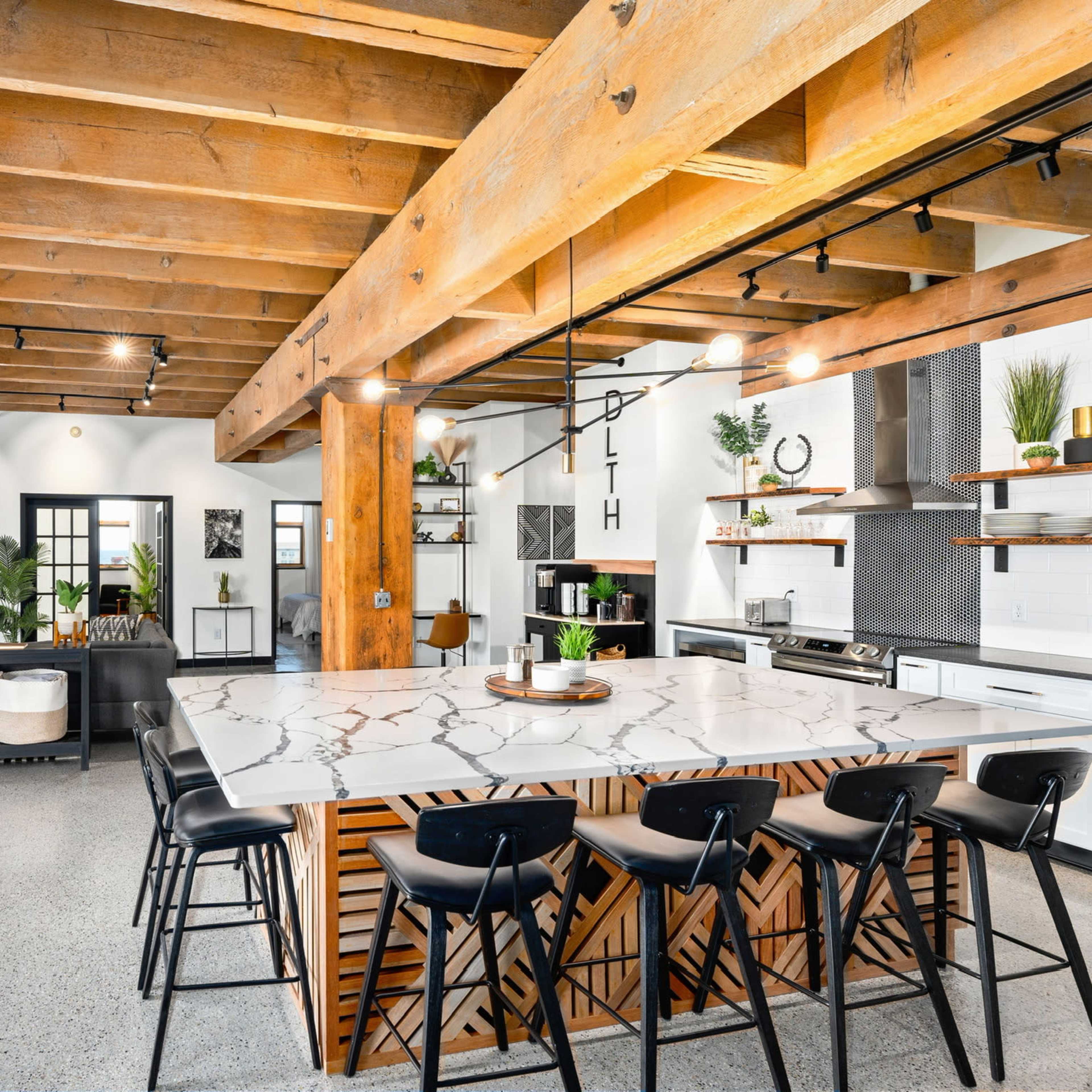 A modern kitchen with a large marble island, black bar stools, wooden beams, and open space leading into a living area with a fireplace and decorated walls.