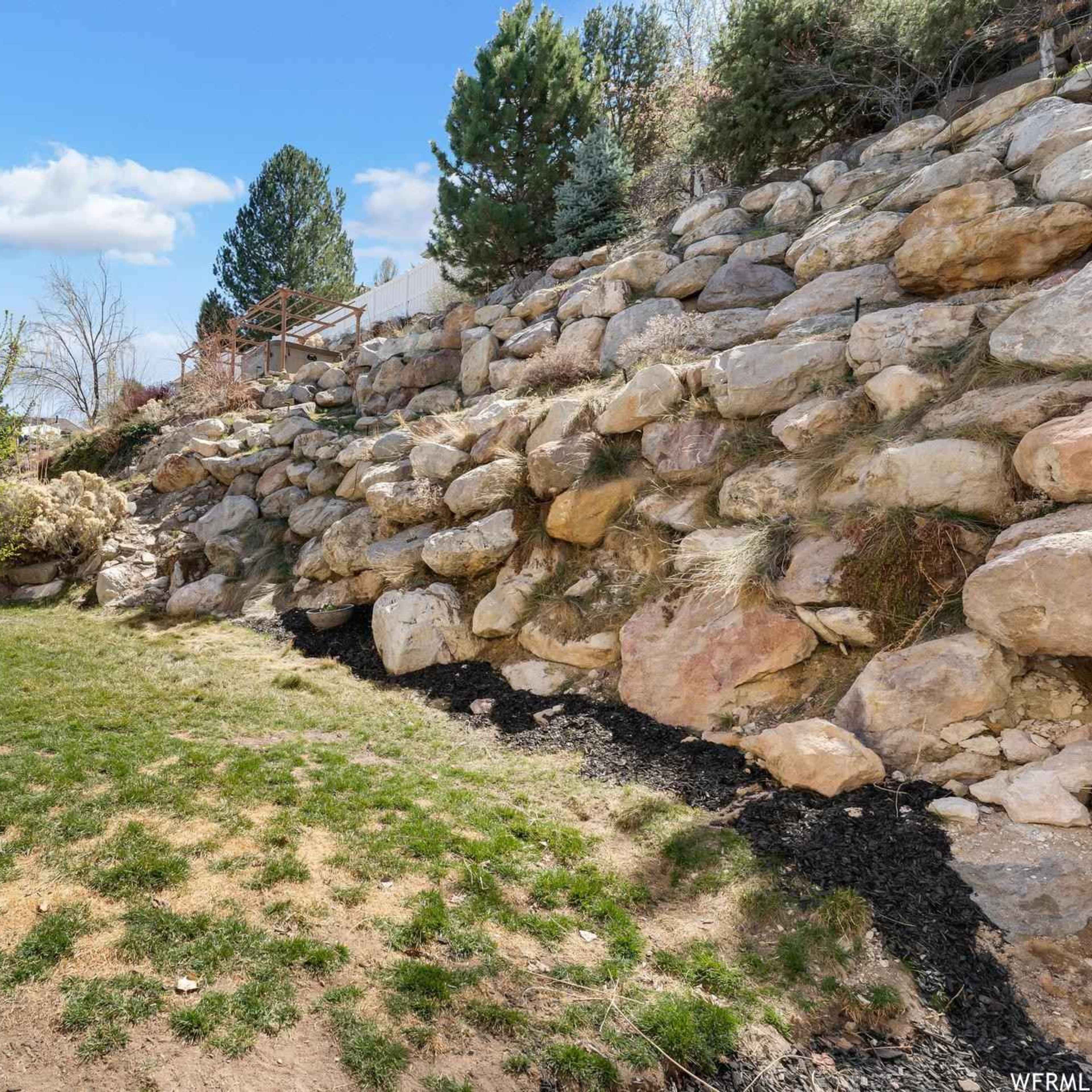A sloped backyard with a rocky hillside, grassy areas, and a house on the left side.