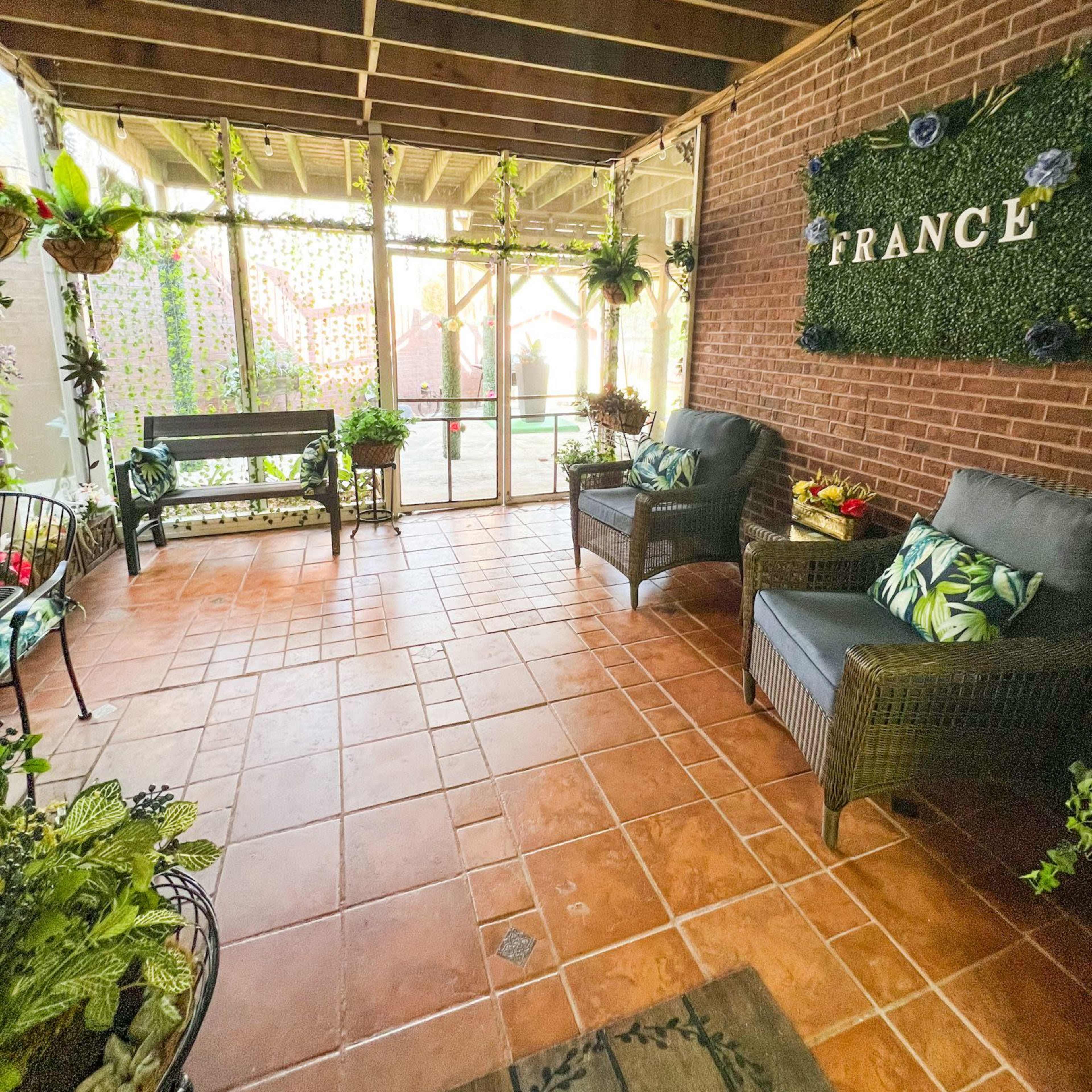 A sunlit indoor space with terracotta tile flooring, featuring wicker seating, potted plants, and a decorative wall that says "FRANCE."