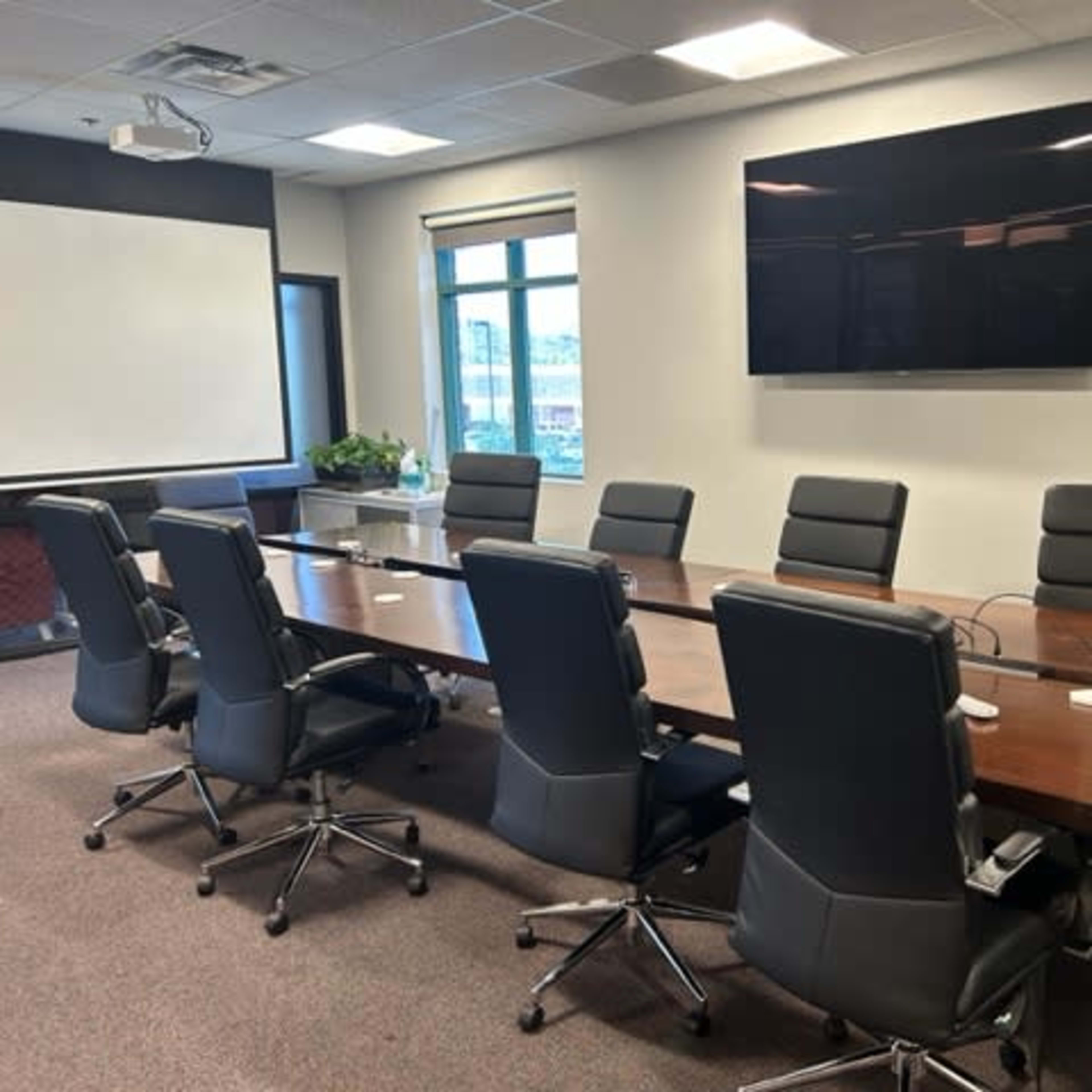 A modern conference room with a long wooden table surrounded by black ergonomic chairs, a wall-mounted screen, and large windows letting in natural light.