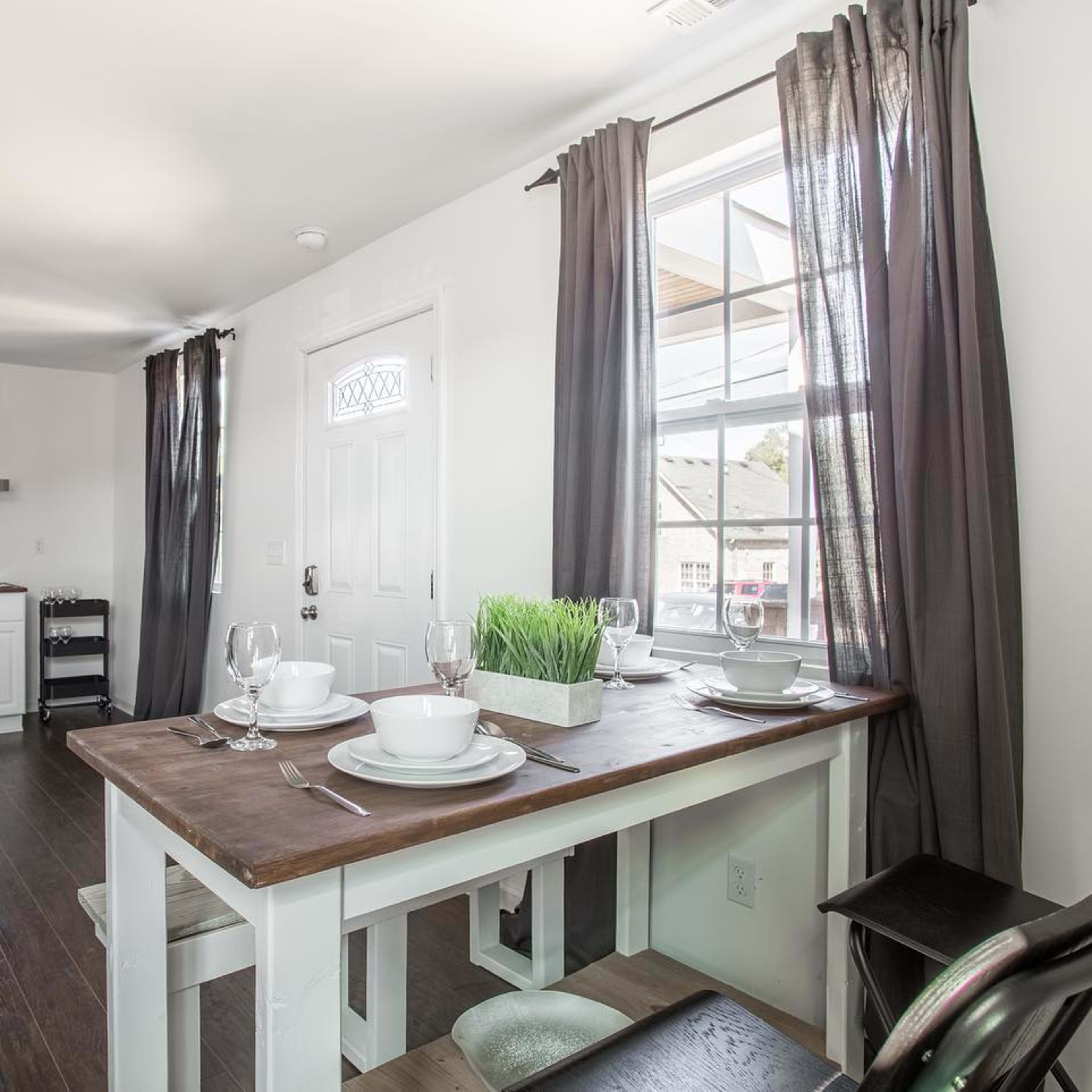 A dining area featuring a wooden table set with dishes and glasses, accompanied by a small kitchen area and windows with dark curtains.
