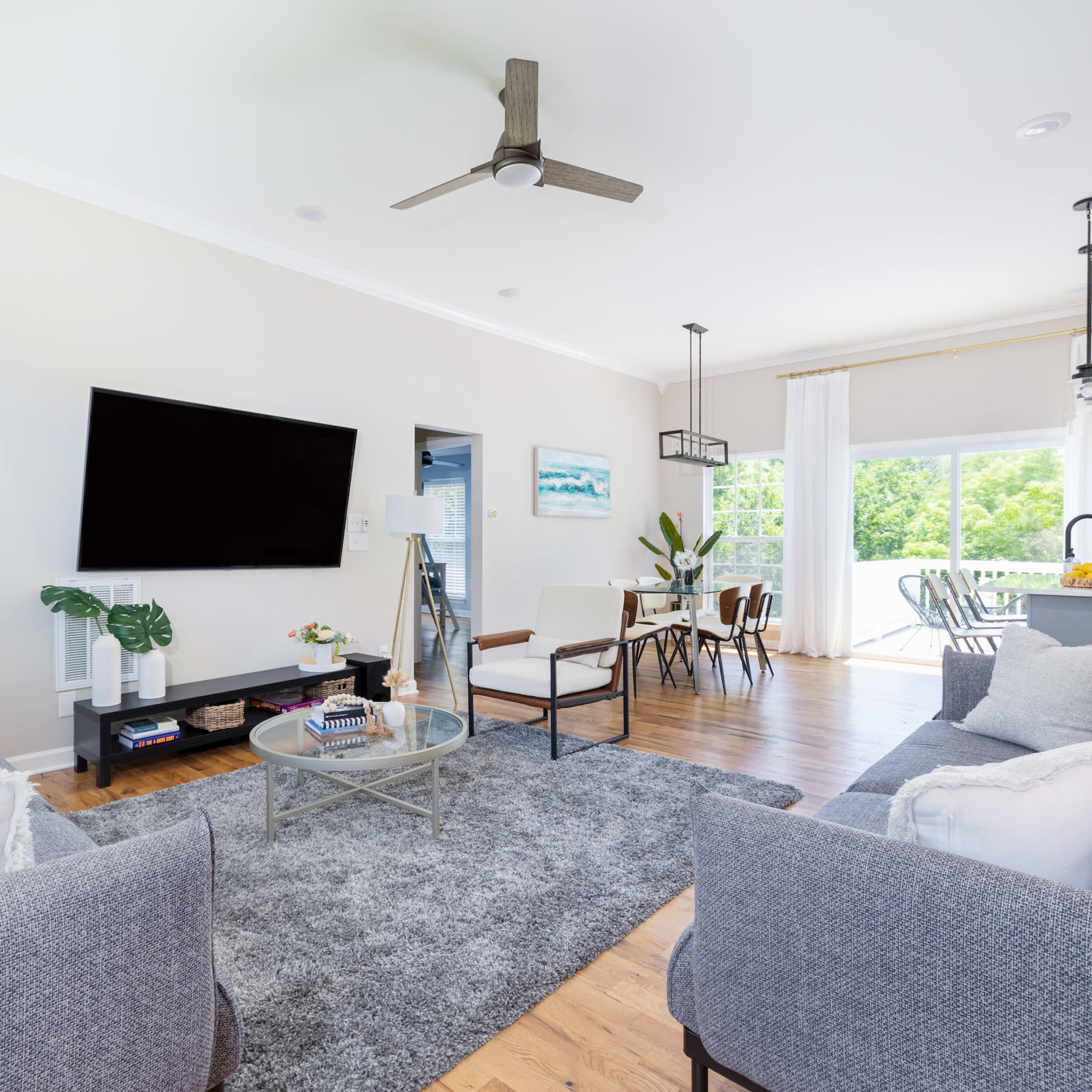 A modern living room with a gray sofa, a coffee table, a wall-mounted television, and a dining area visible through a large window.