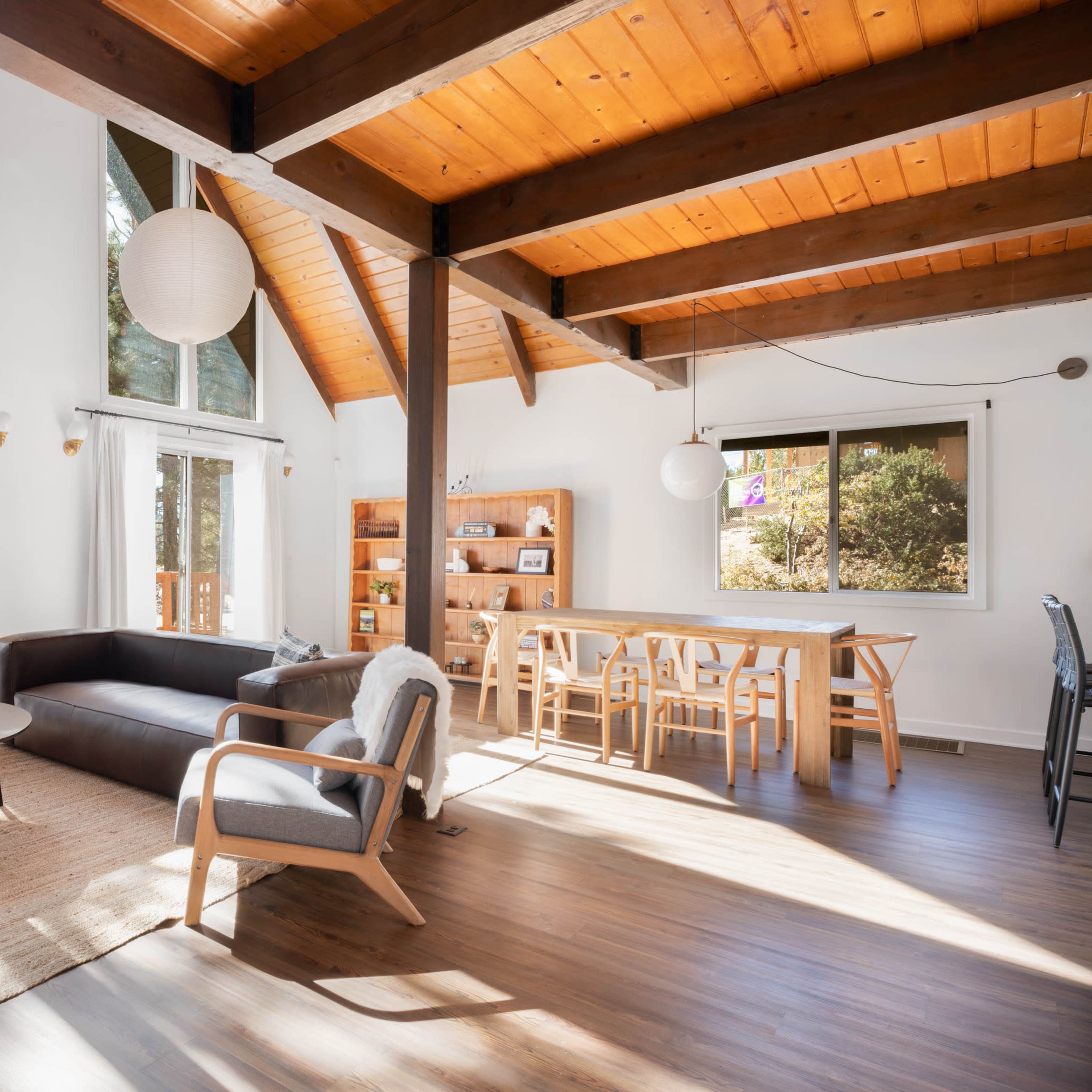 A bright, open living space with a sectional couch, dining area, and large windows showcasing a view of trees outside.
