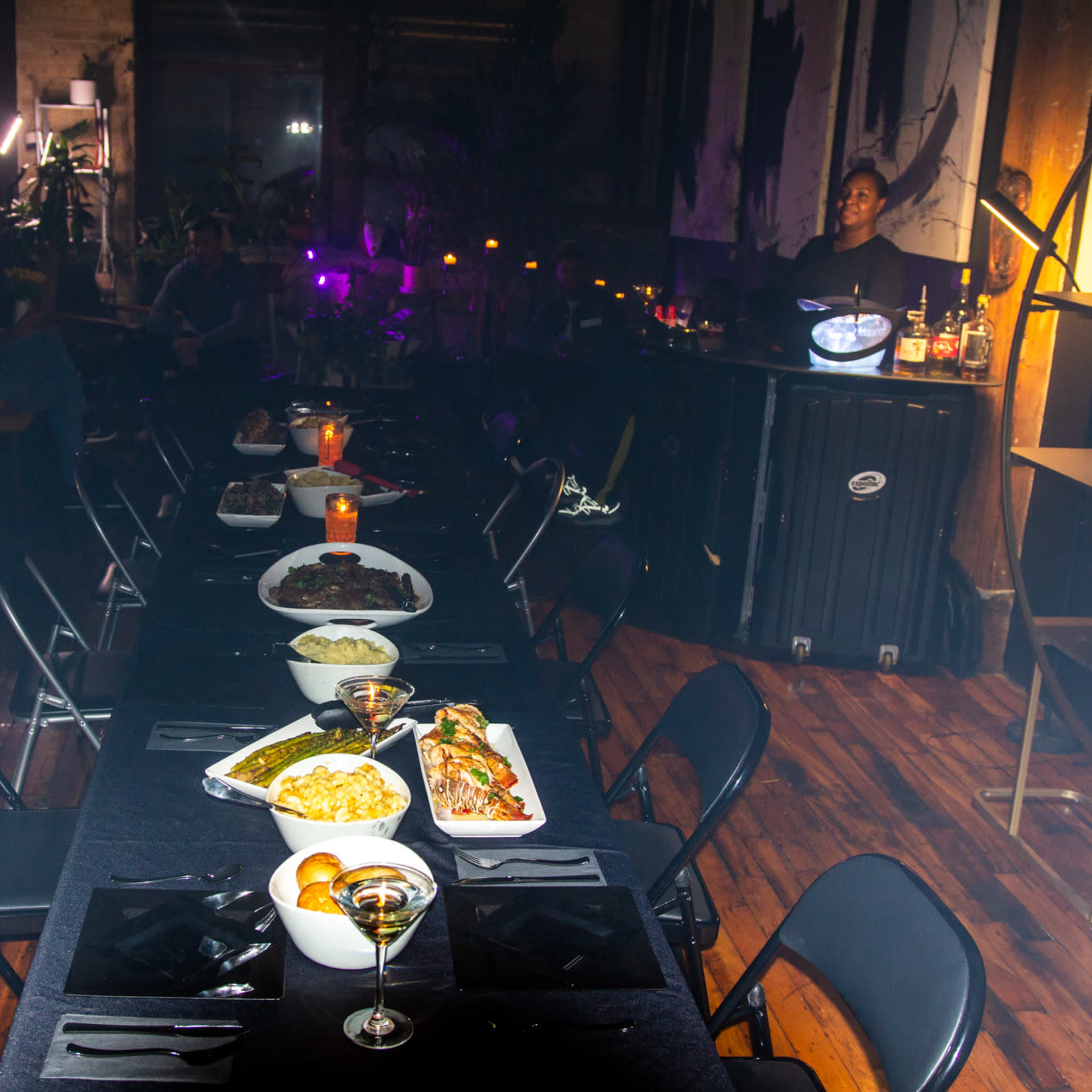 A long dining table is set with various dishes and drinks in a dimly lit room featuring a bar and people in the background.