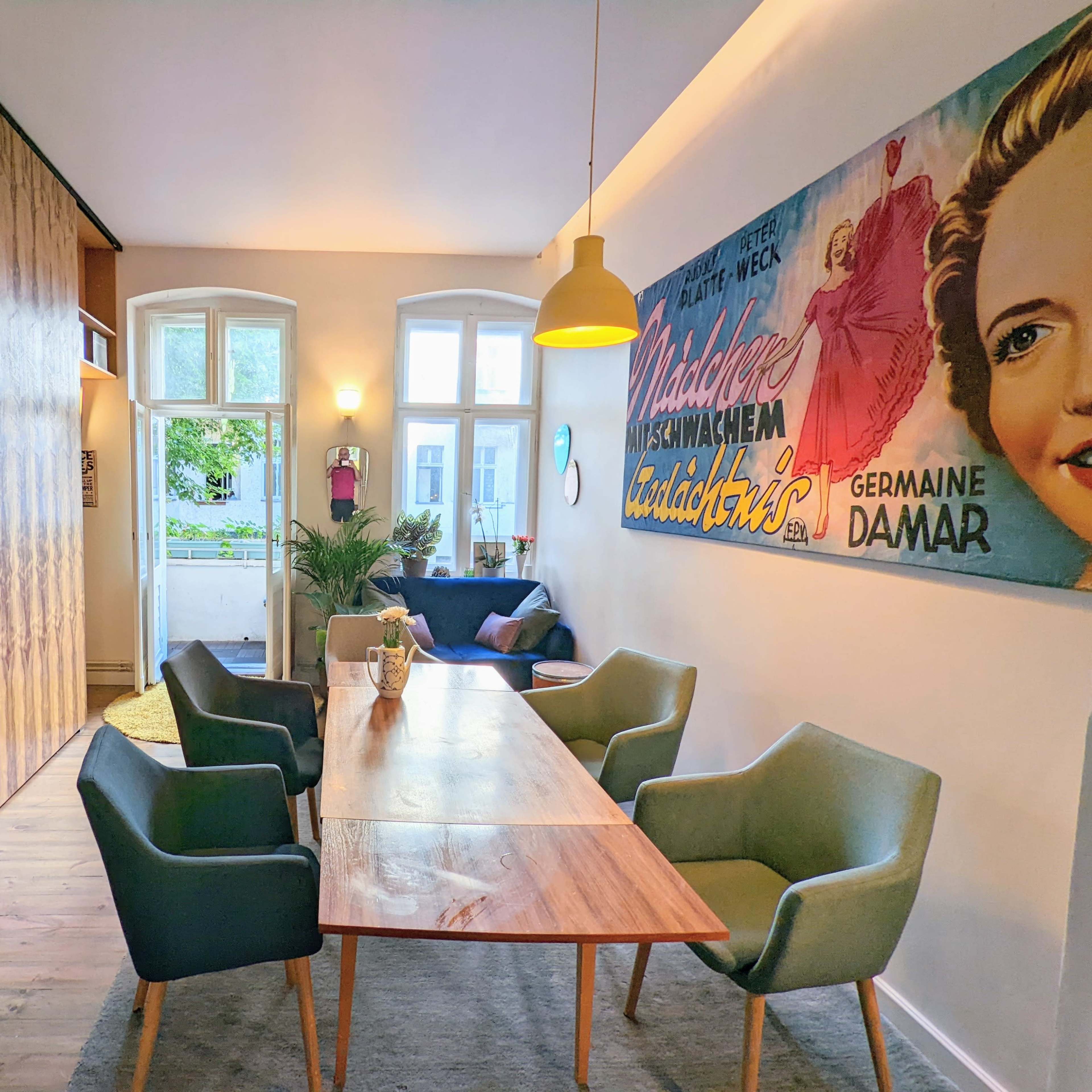 A modern living space with a wooden dining table surrounded by four chairs, a bookshelf filled with plants and books, and a vibrant wall adorned with a vintage poster.