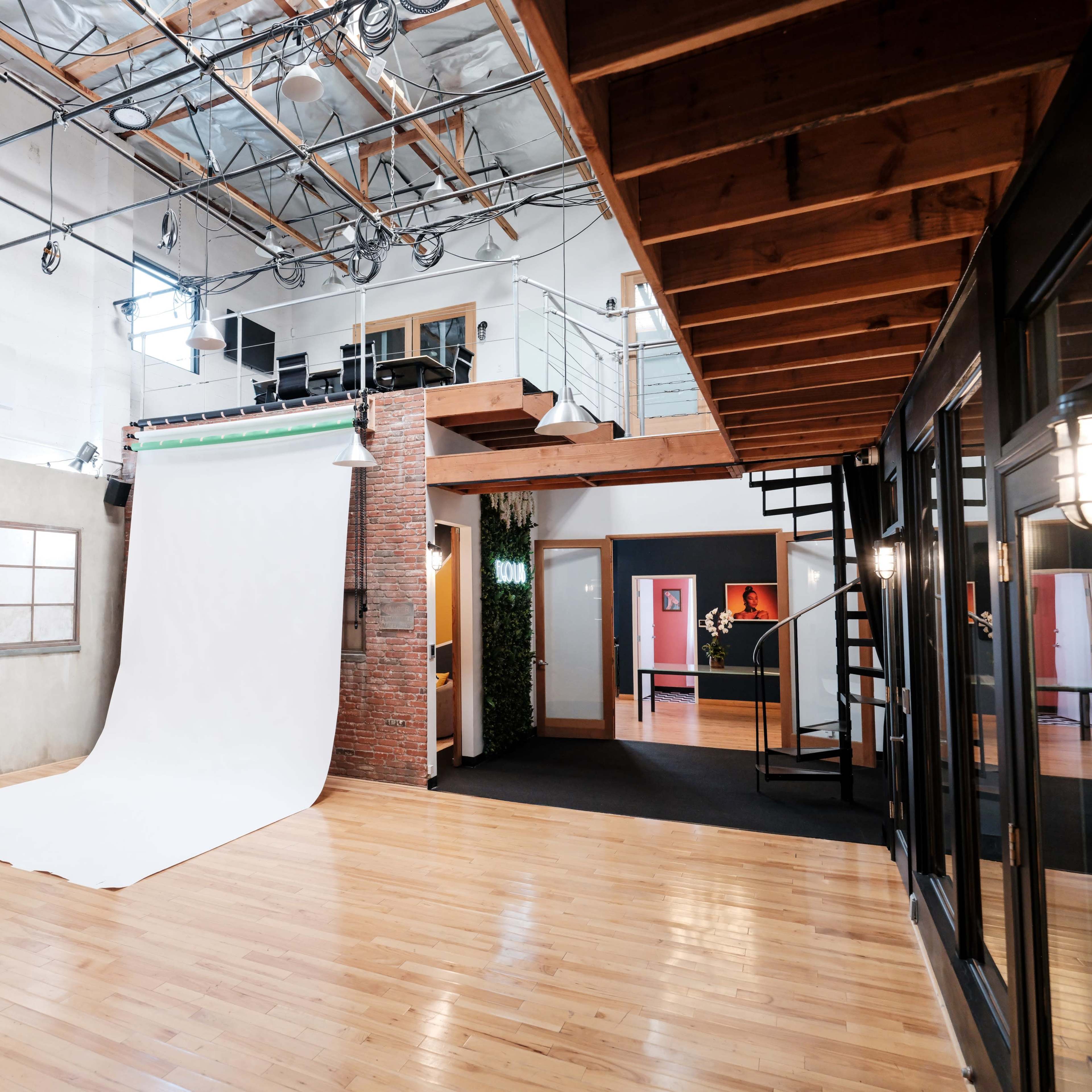 A spacious photography studio with a large white backdrop, wooden floors, and a modern design featuring a lofted area with seating and multiple entrances.
