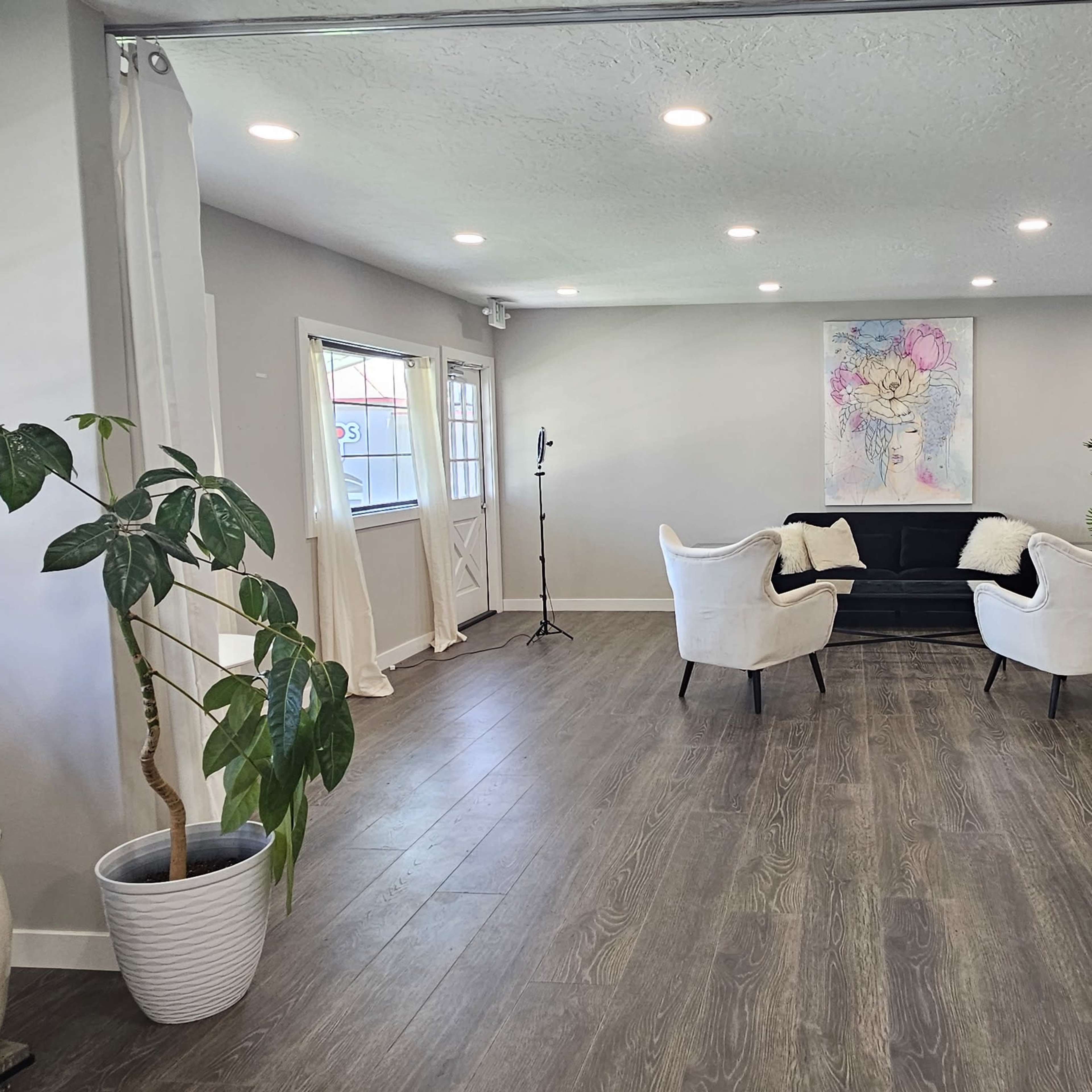 A modern, open room with a seating area featuring two white chairs and a black couch, along with plants and a desk in a well-lit environment.