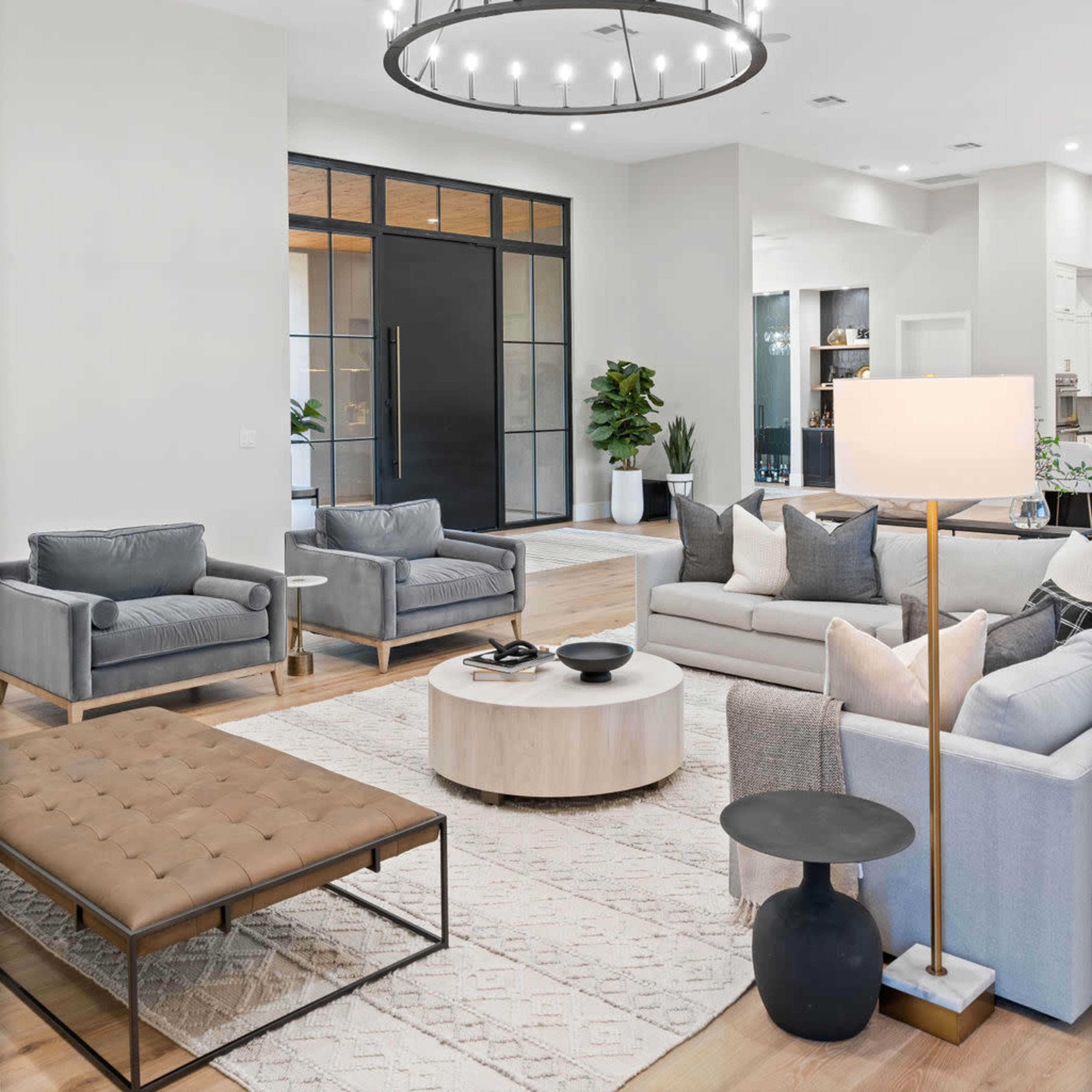 A modern living room features gray upholstered furniture, a round coffee table, and a large chandelier above, with glass doors leading to another area.