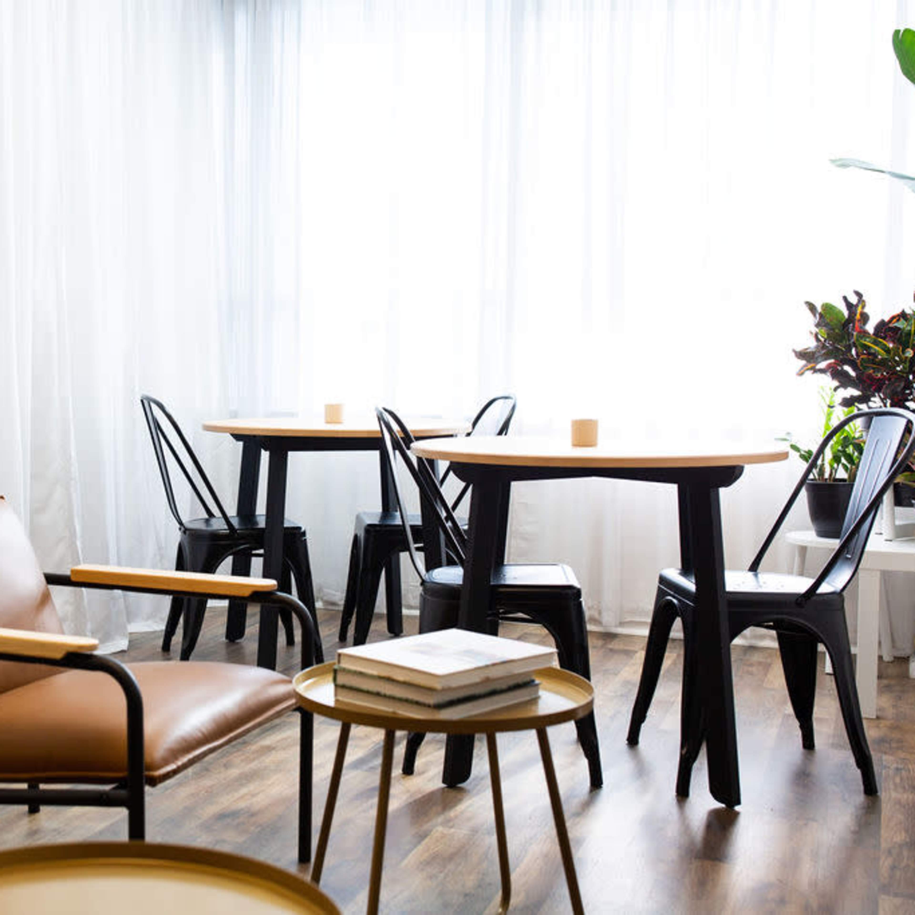 A bright, minimalist room featuring brown leather chairs, black metal tables, and large windows dressed with sheer white curtains.
