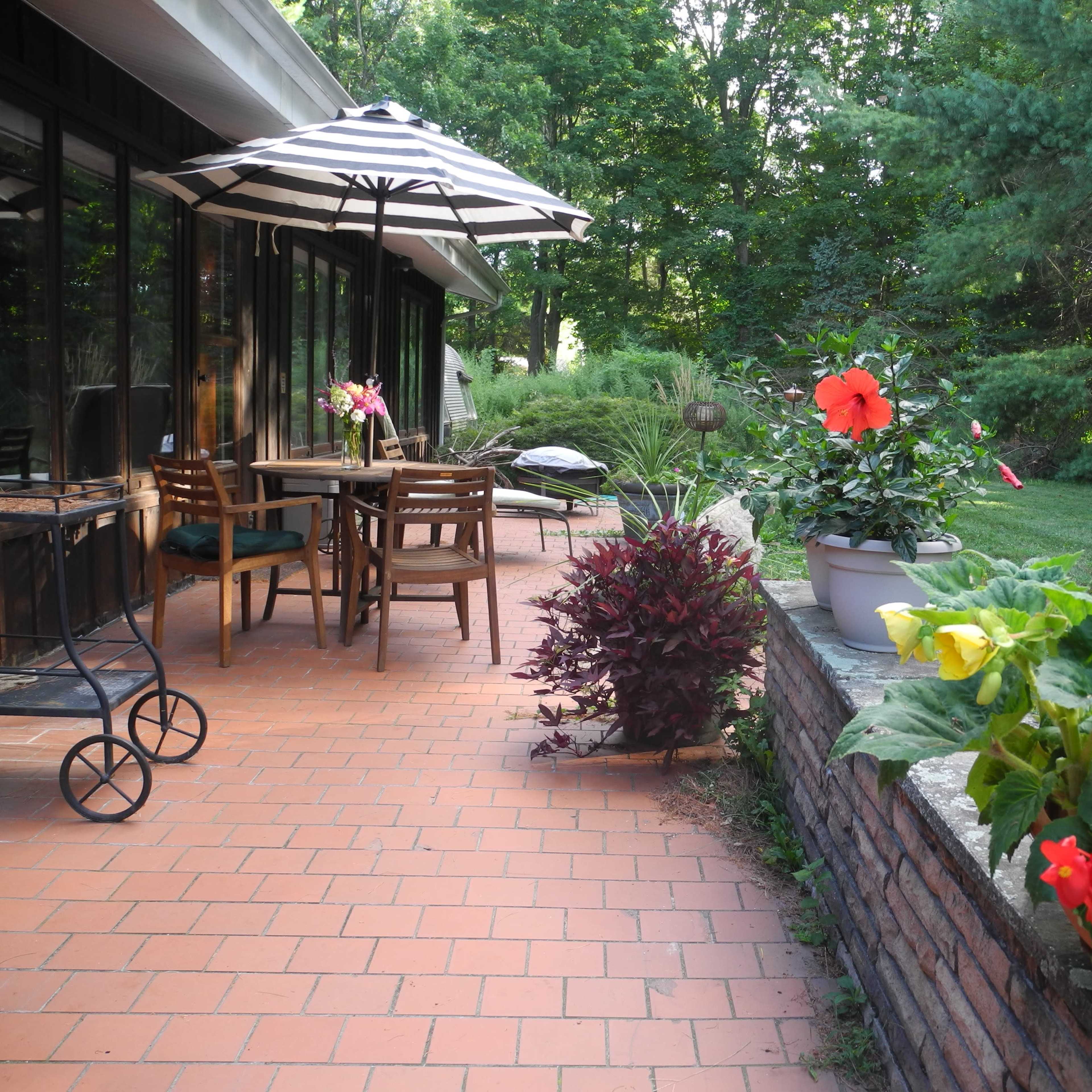A patio with a table and chairs under a striped umbrella, surrounded by potted plants and flowers.