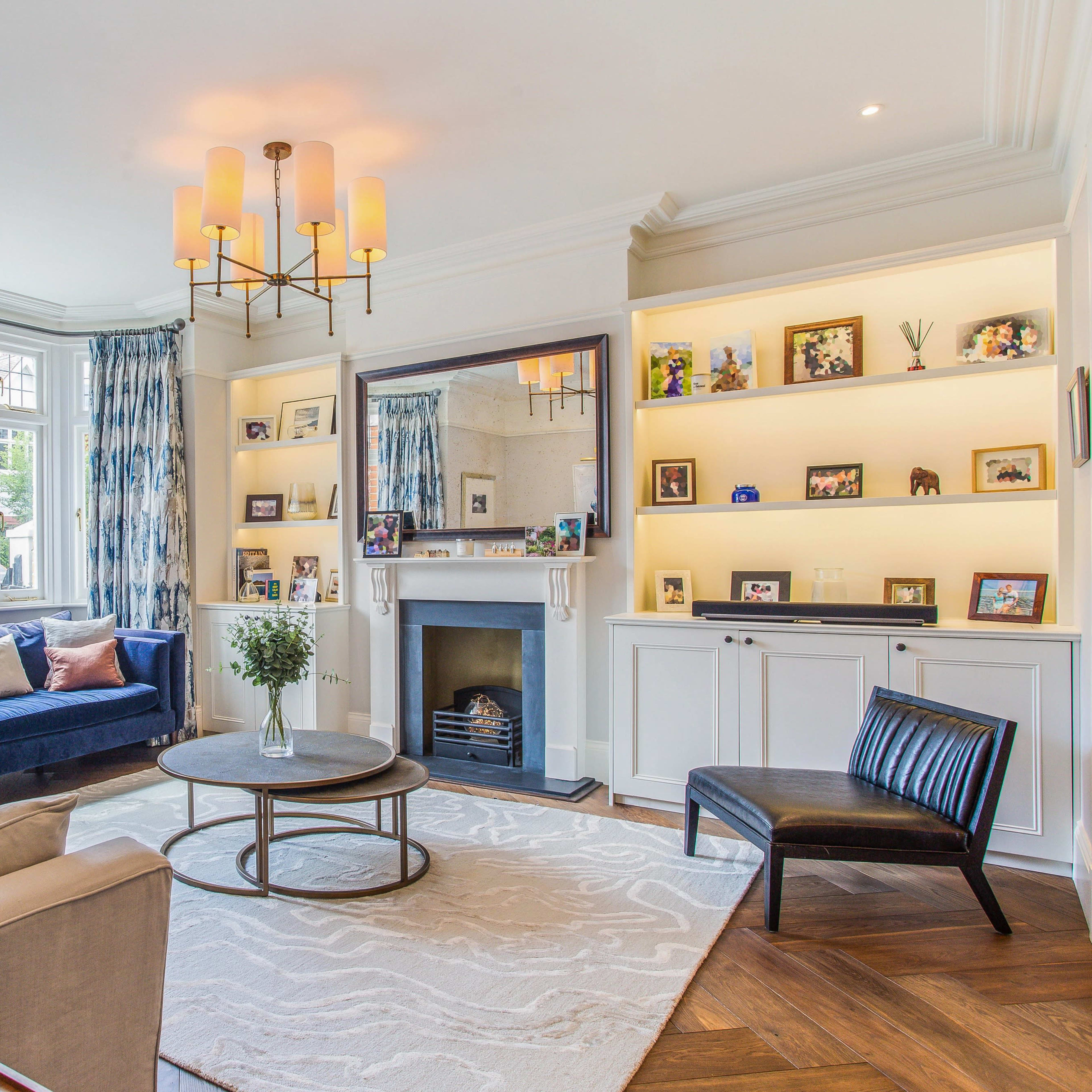 The living room features a blue sofa, a round coffee table, and a fireplace, all arranged around a large window and surrounded by decorative shelving.