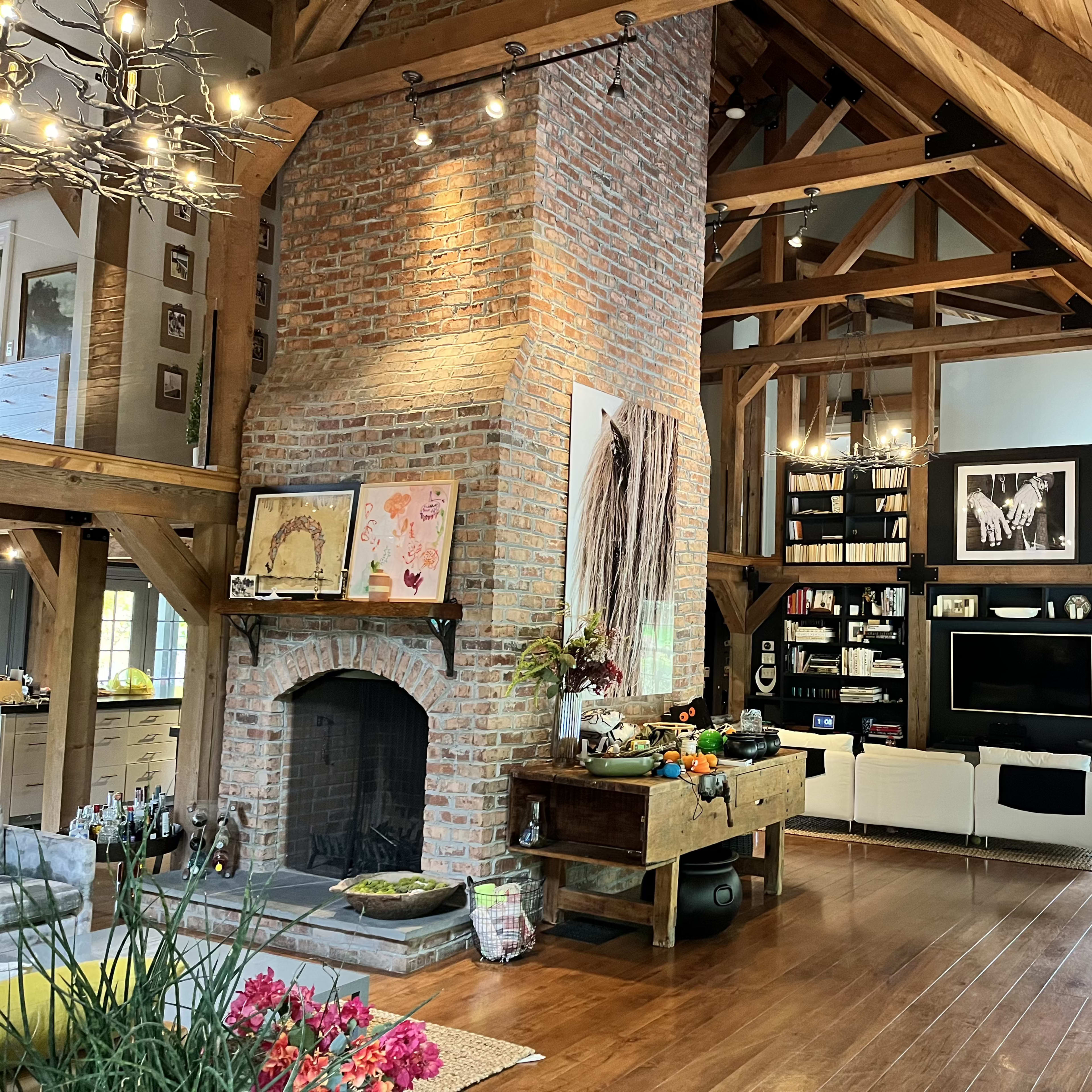 A spacious living room features a tall brick fireplace, wooden beams, and modern furnishings, illuminated by overhead lighting.