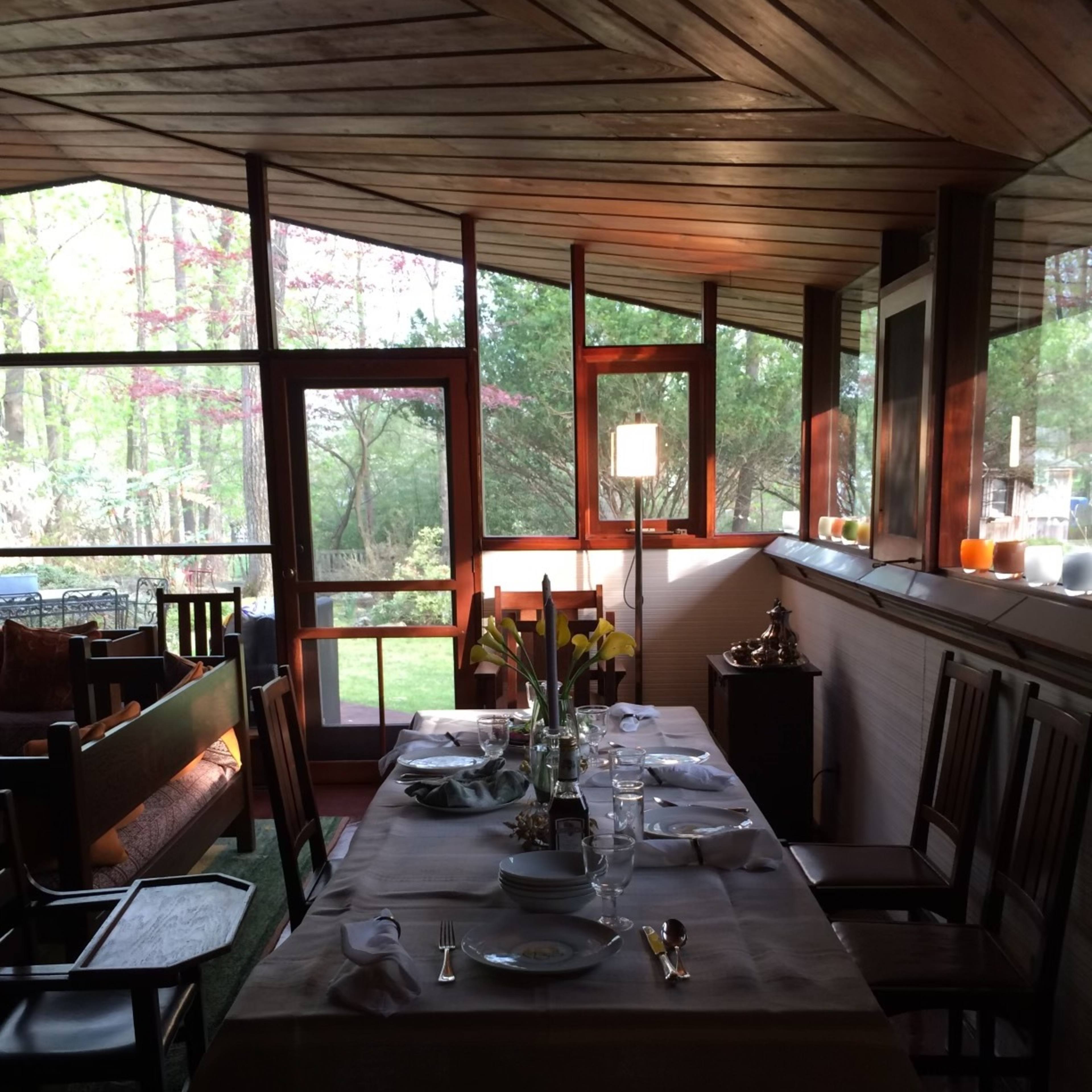 A dining table is set with plates and glasses in a sunlit room surrounded by large windows overlooking a garden.