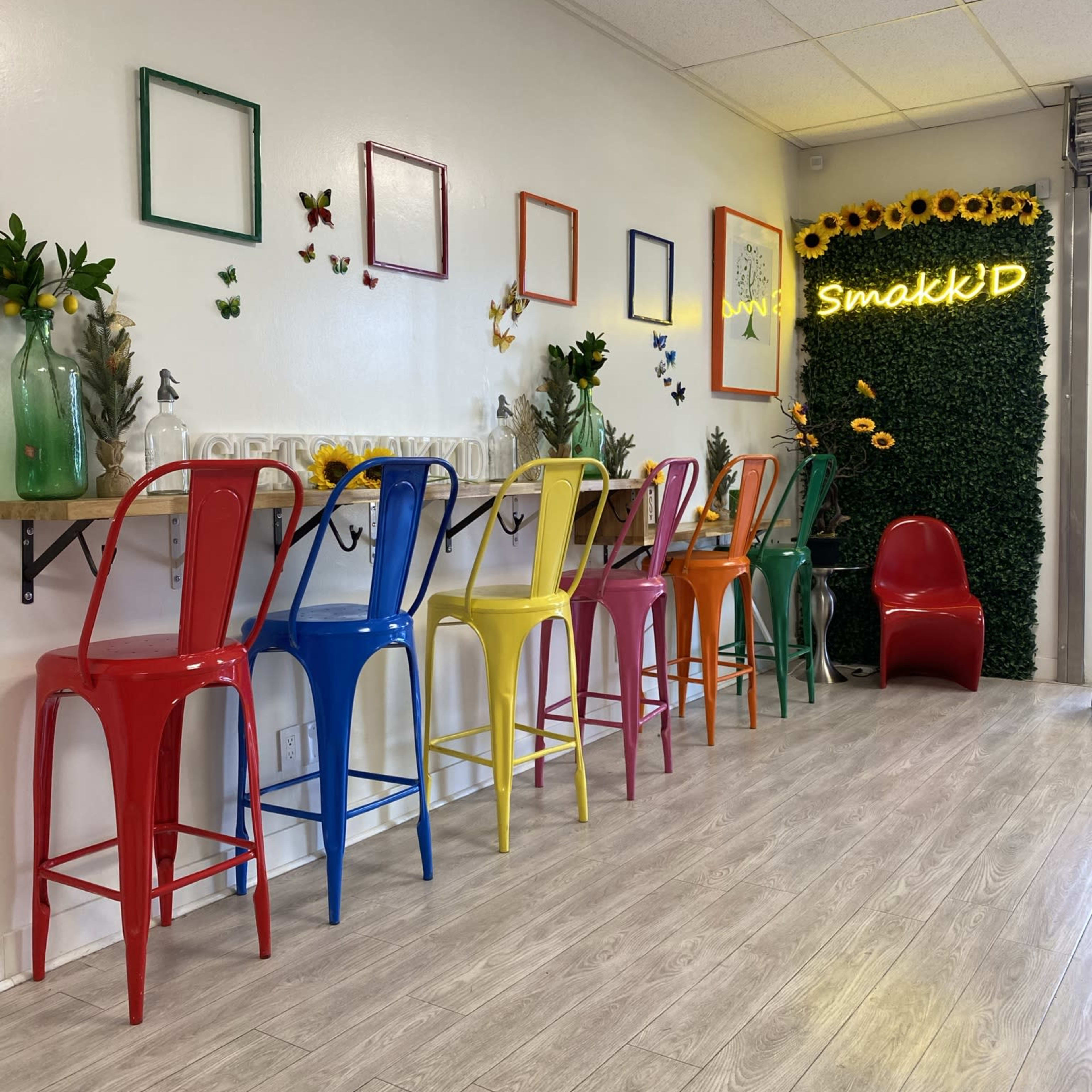 A brightly colored dining area with metal chairs in red, blue, yellow, and green arranged around a long wooden table, decorated with plants and wall art.