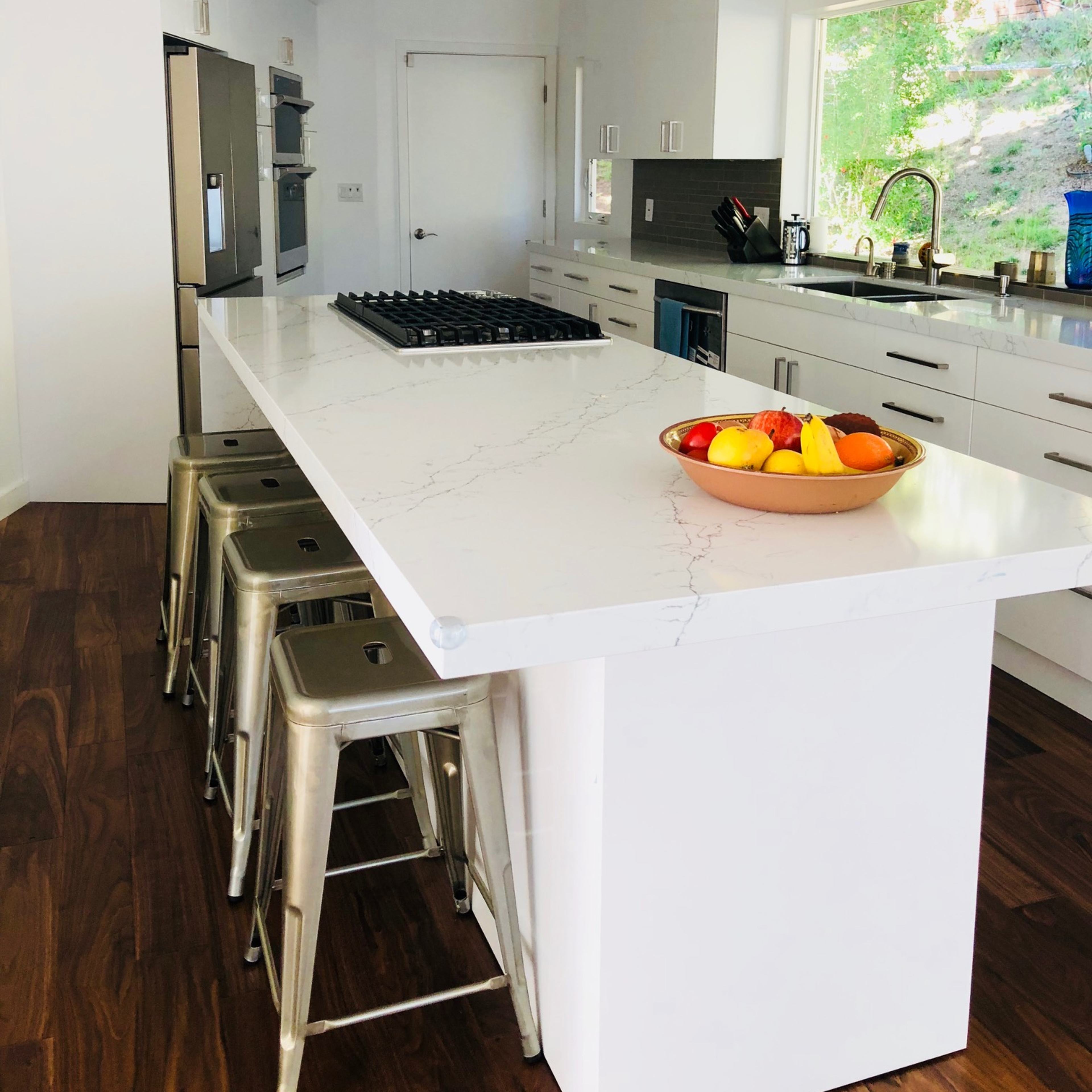 A modern kitchen features a large white island with a marble top and metal bar stools, surrounded by sleek cabinetry and a view of greenery outside.