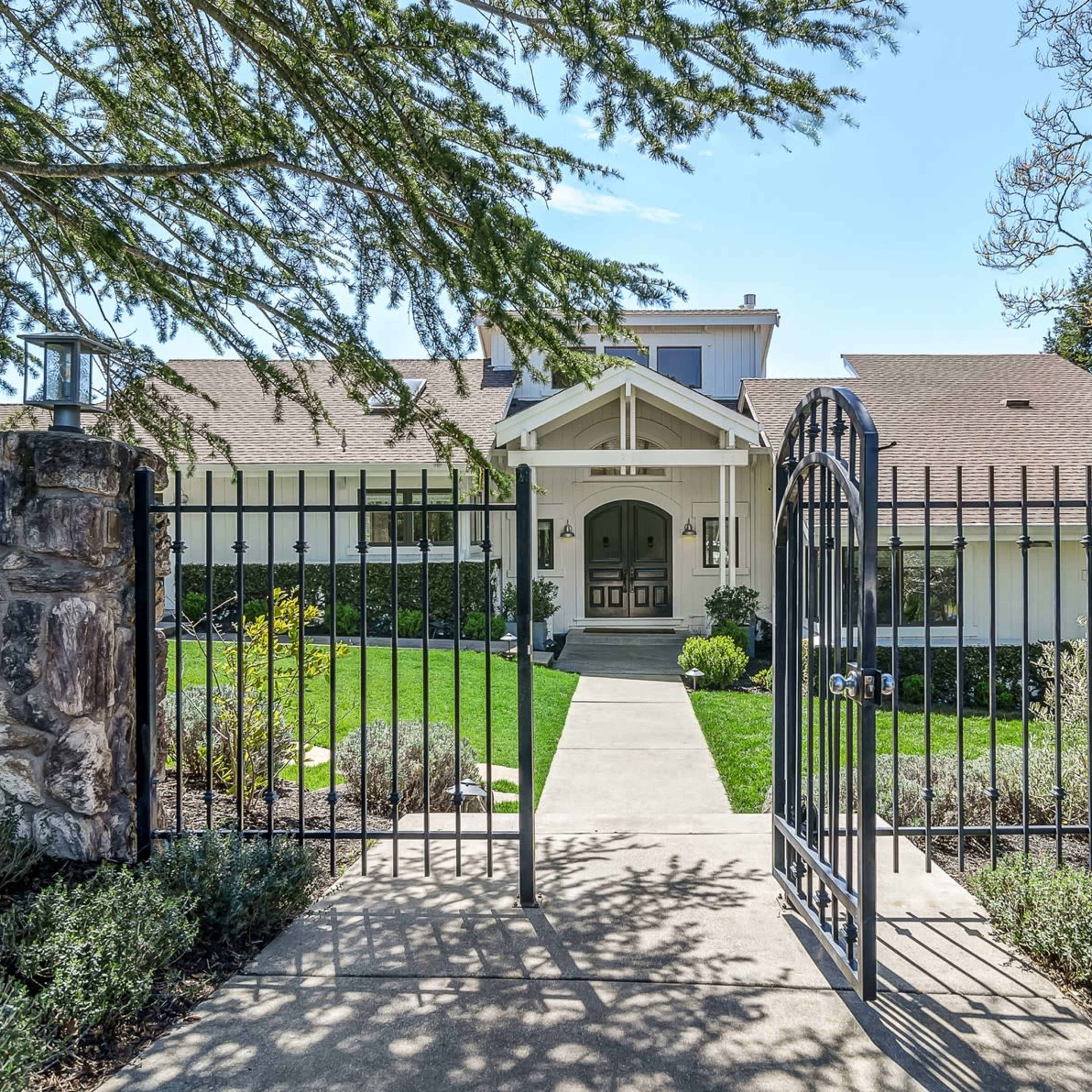 A stone gate opens to a well-maintained pathway leading to a modern house with a front porch and manicured landscaping.