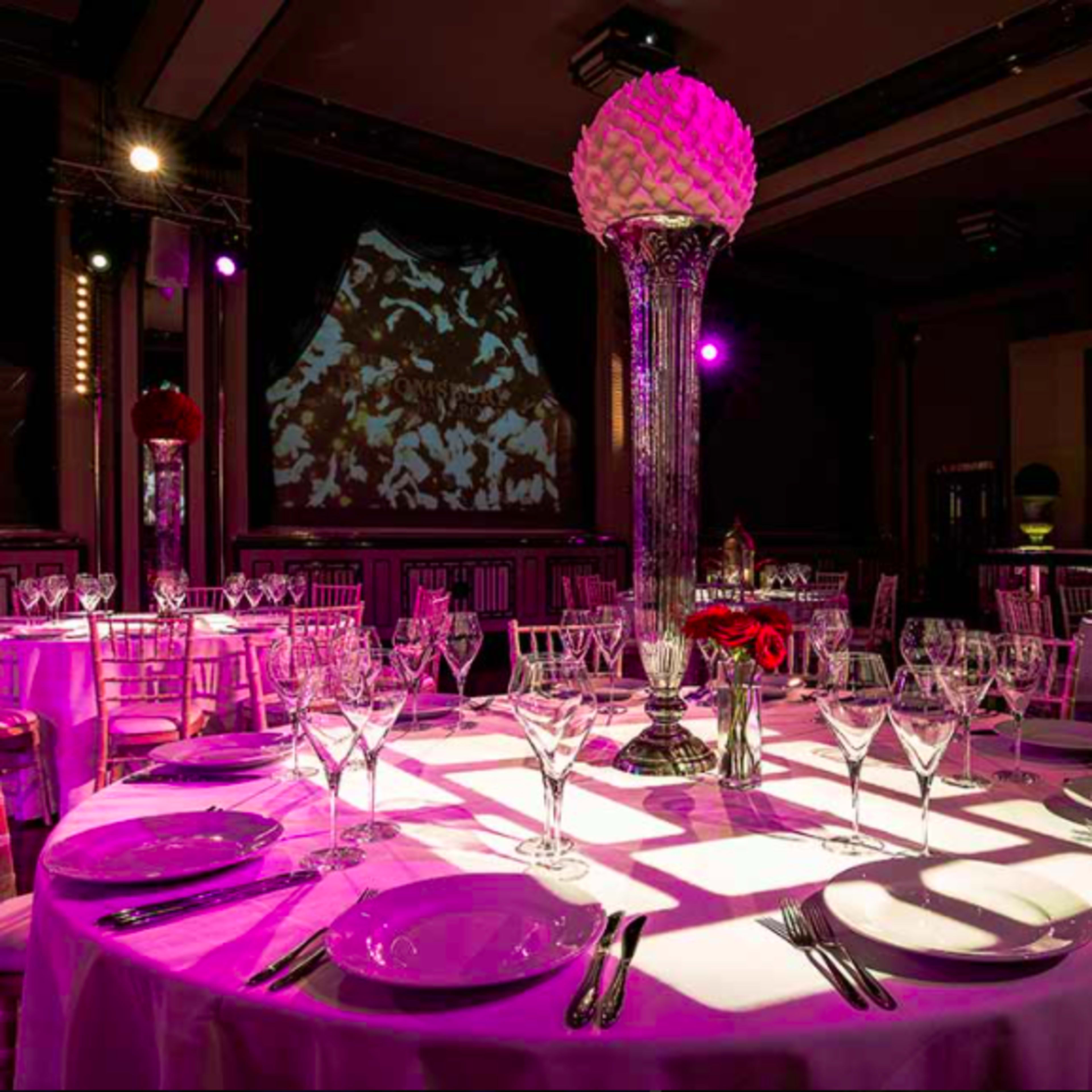 A beautifully set dining table with elegant glassware and silverware, illuminated by purple lighting in a luxurious banquet hall.