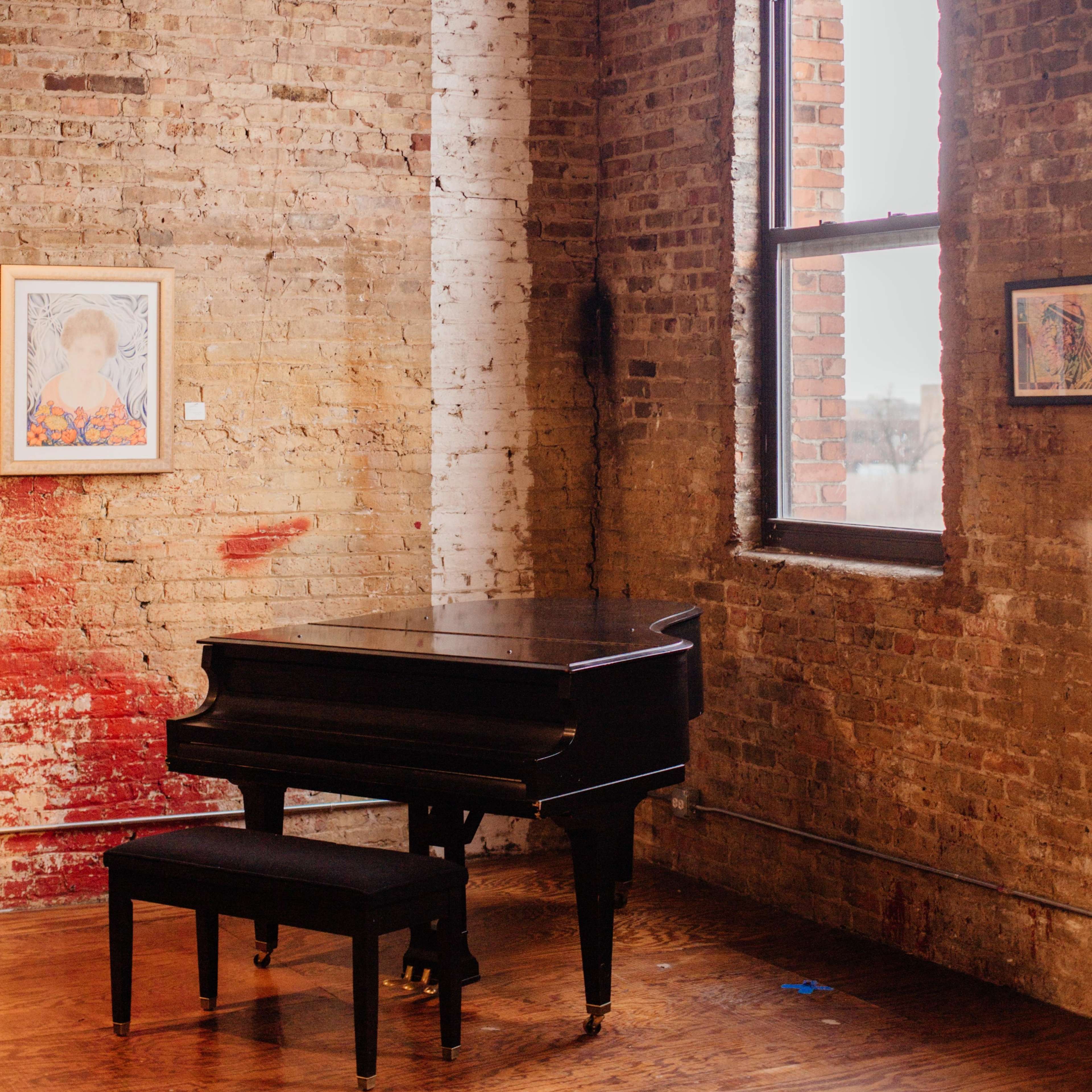 A black grand piano sits next to a window in a room with exposed brick walls and framed artwork.