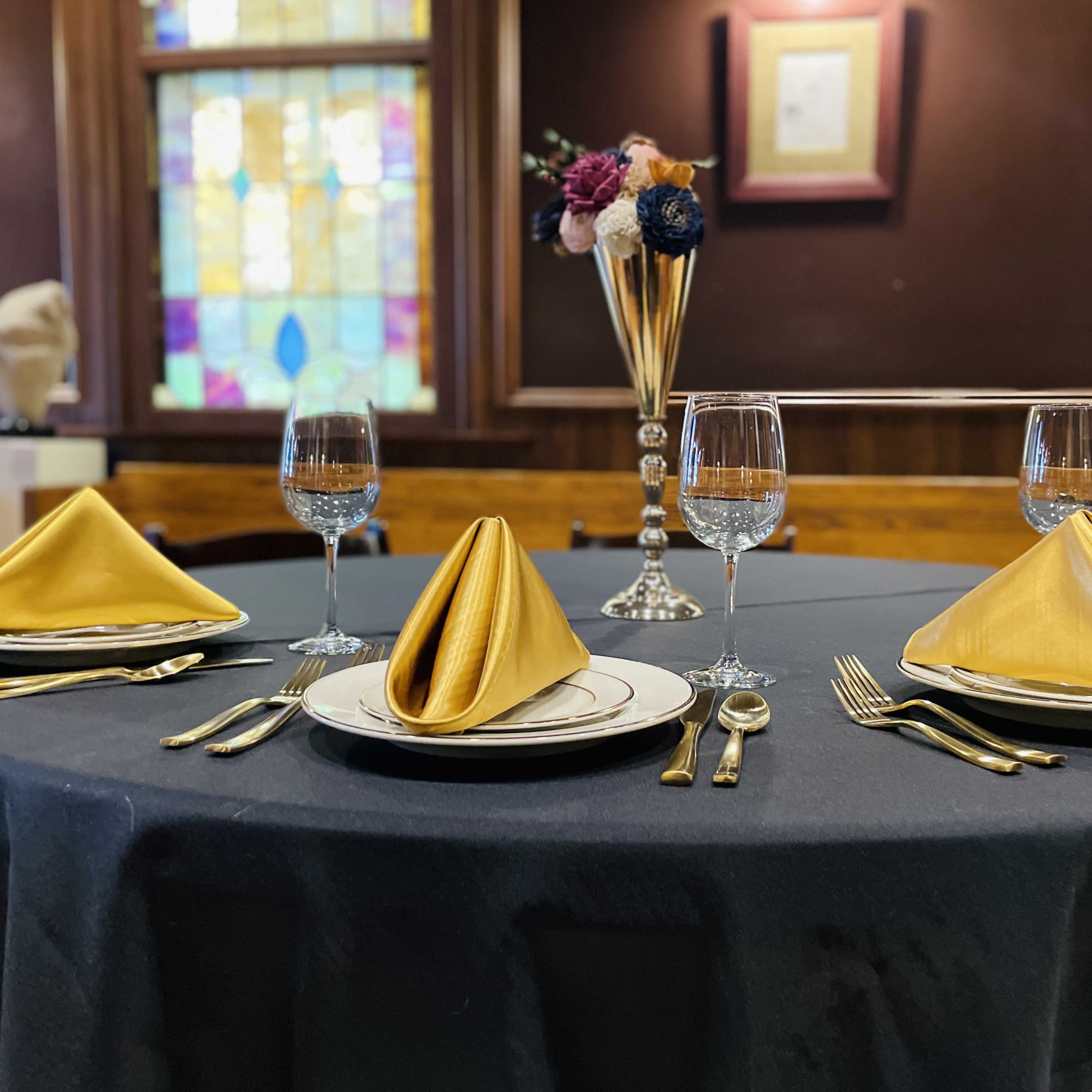 A banquet table set with gold napkins, elegant dishware, glassware, and a floral centerpiece, against a backdrop of stained glass windows.