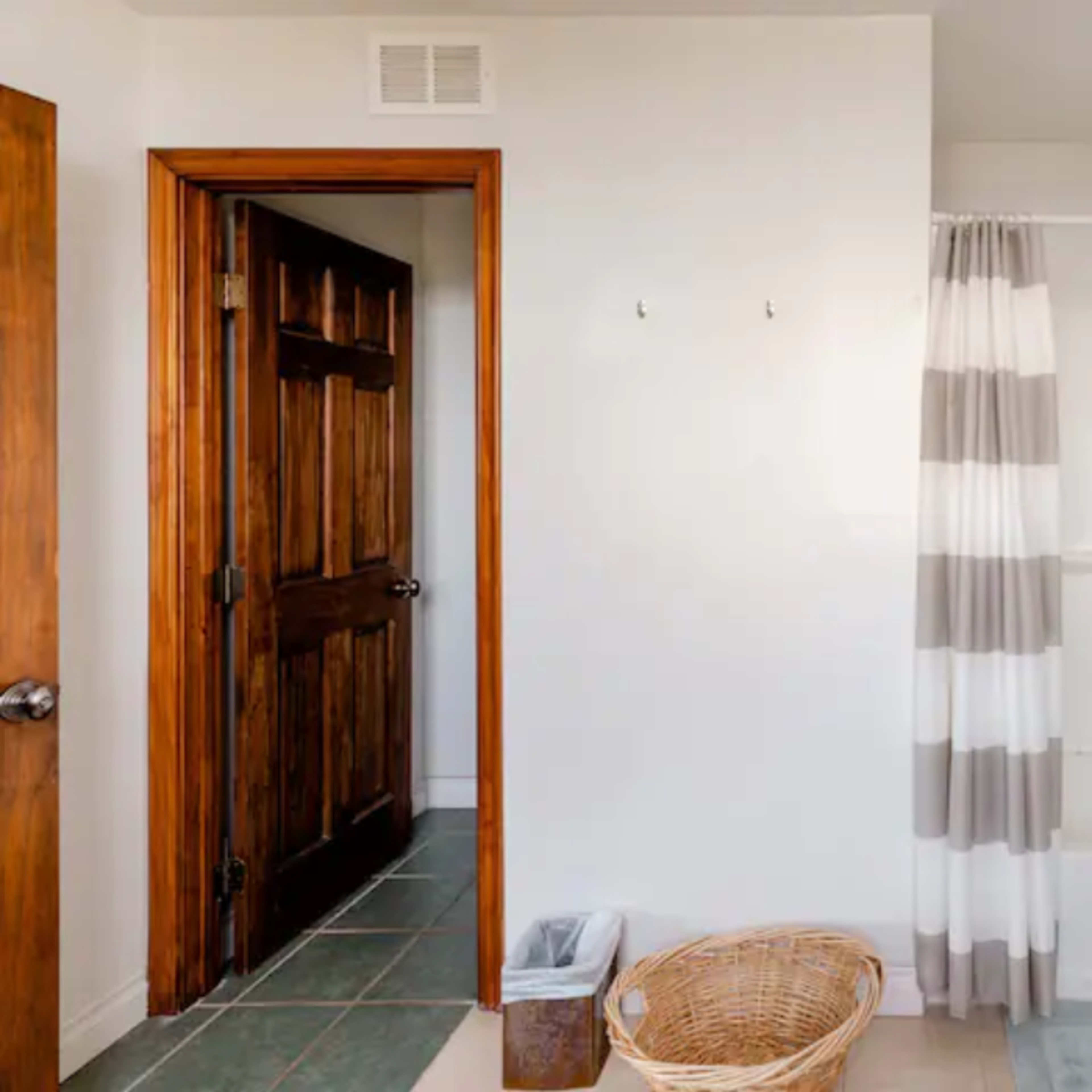 The bathroom features a shower area with a striped curtain, a wooden door, a laundry basket, and a small trash can.