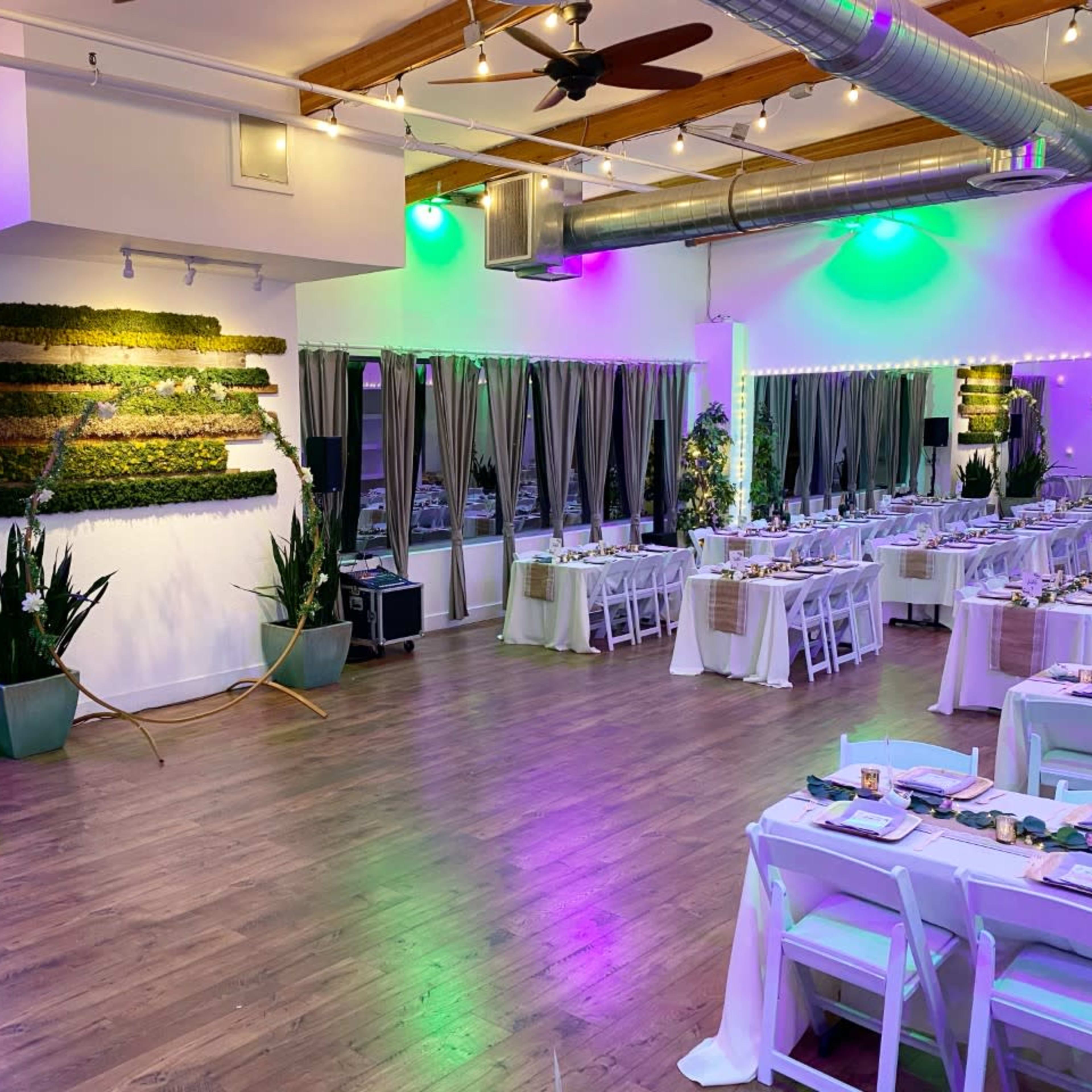 A spacious event room is softly illuminated with green and purple lighting, featuring dining tables arranged neatly with white chairs and a green moss wall as a backdrop.