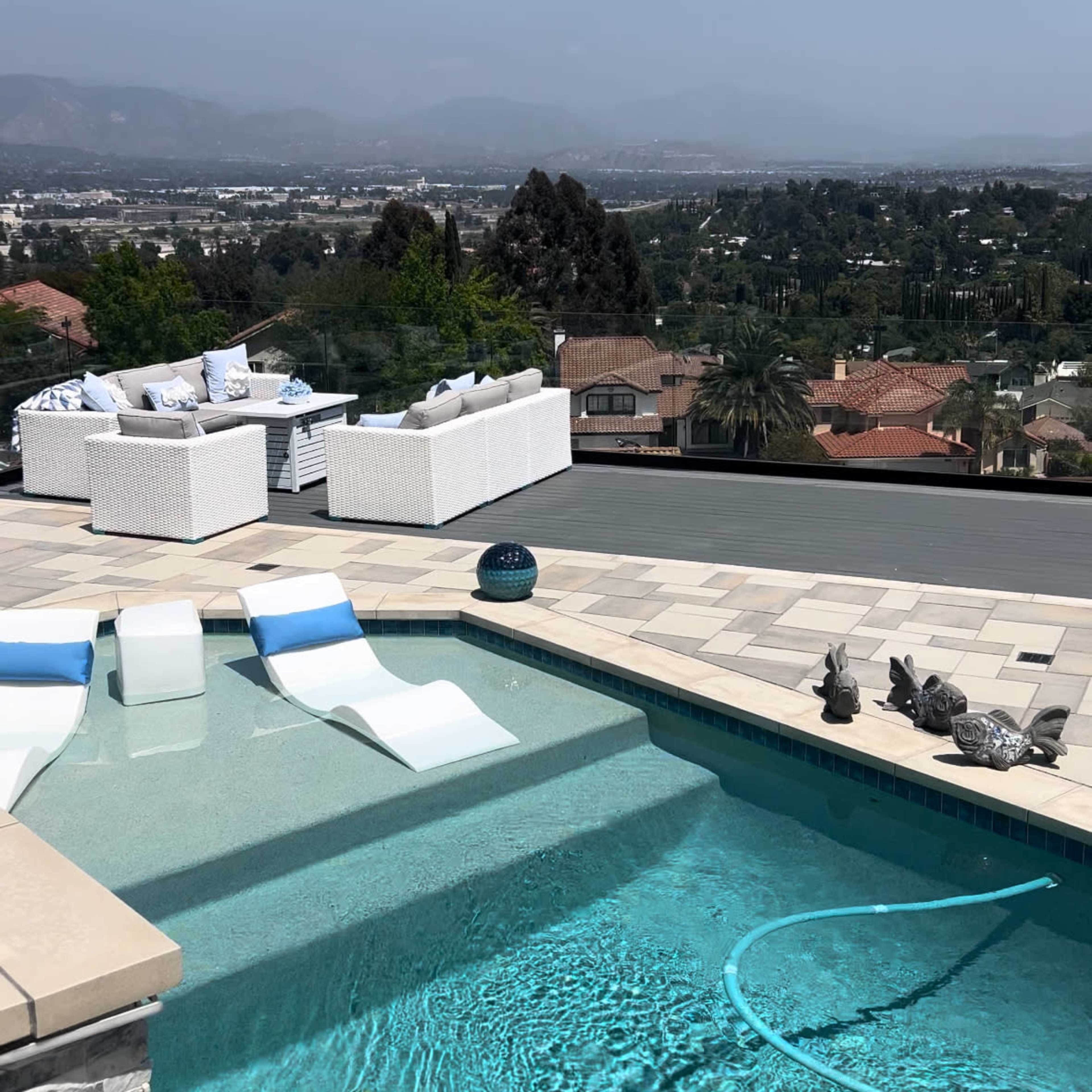 A swimming pool with white lounge chairs is situated on a terrace overlooking a suburban landscape.