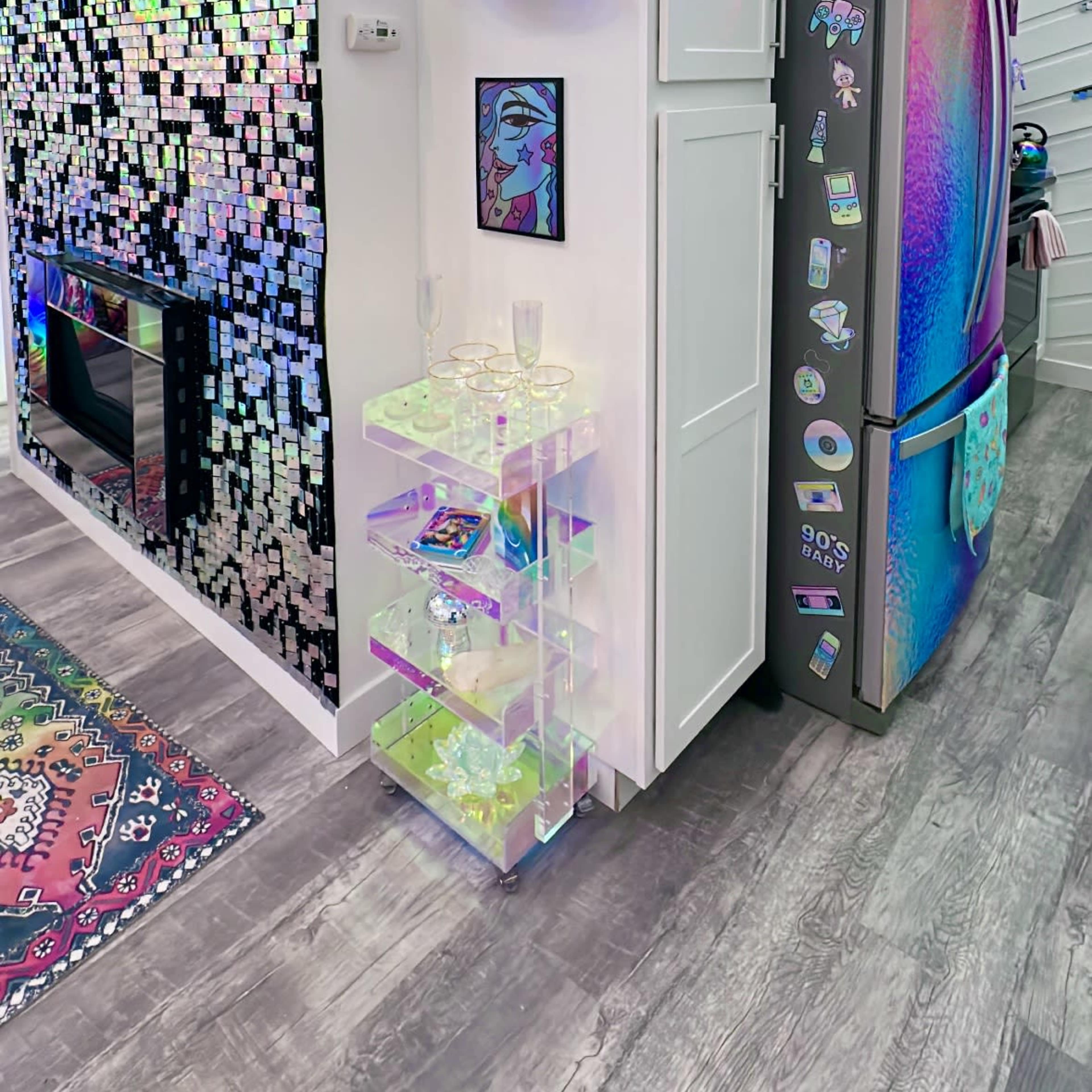 A modern kitchen corner featuring a refrigerator with a reflective, colorful surface, a wall decorated with sequined tiles, and a small transparent shelving unit displaying decorative items.