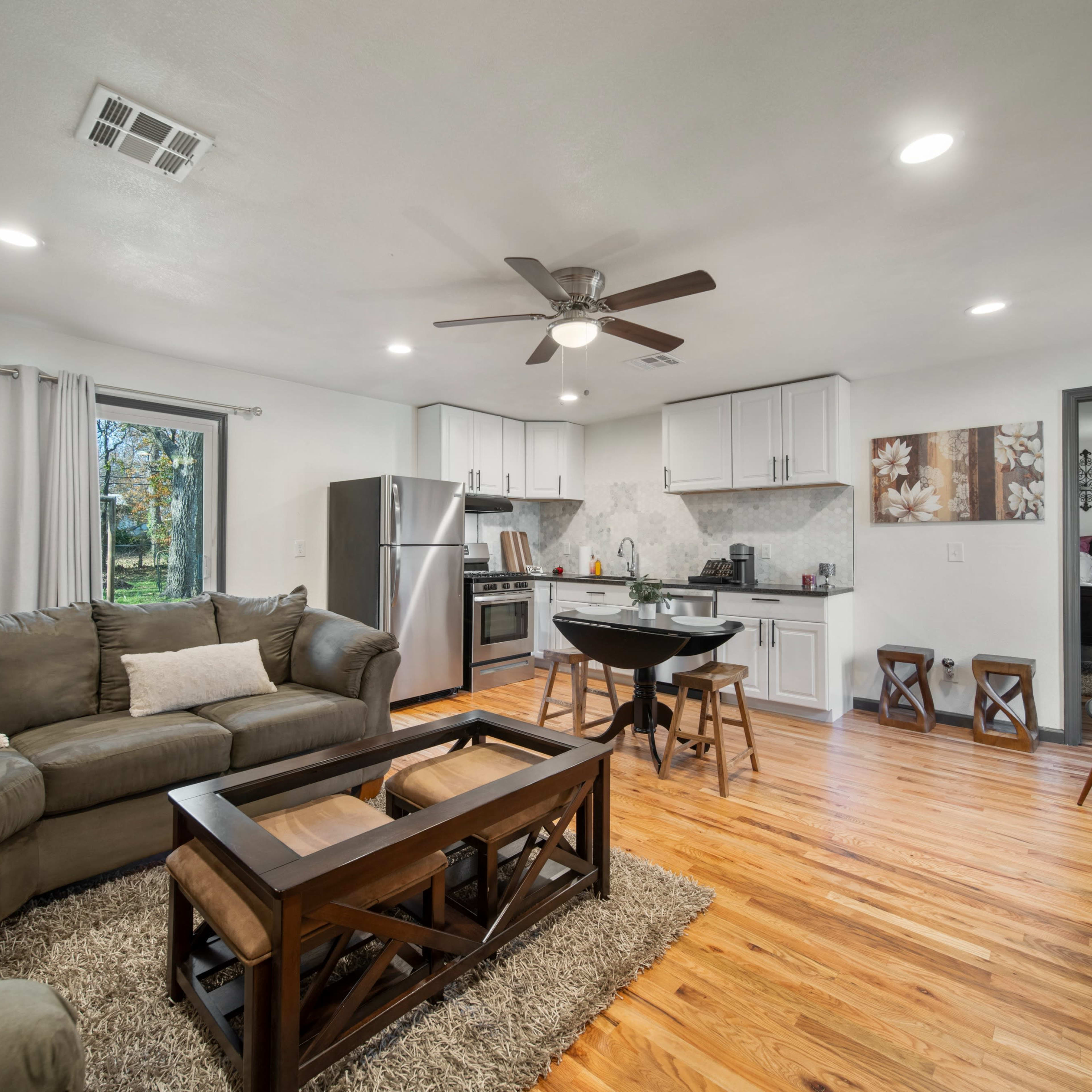 A modern living room features a gray sectional sofa, a wooden coffee table, and a dining area with a round table, surrounded by large windows and wooden flooring.