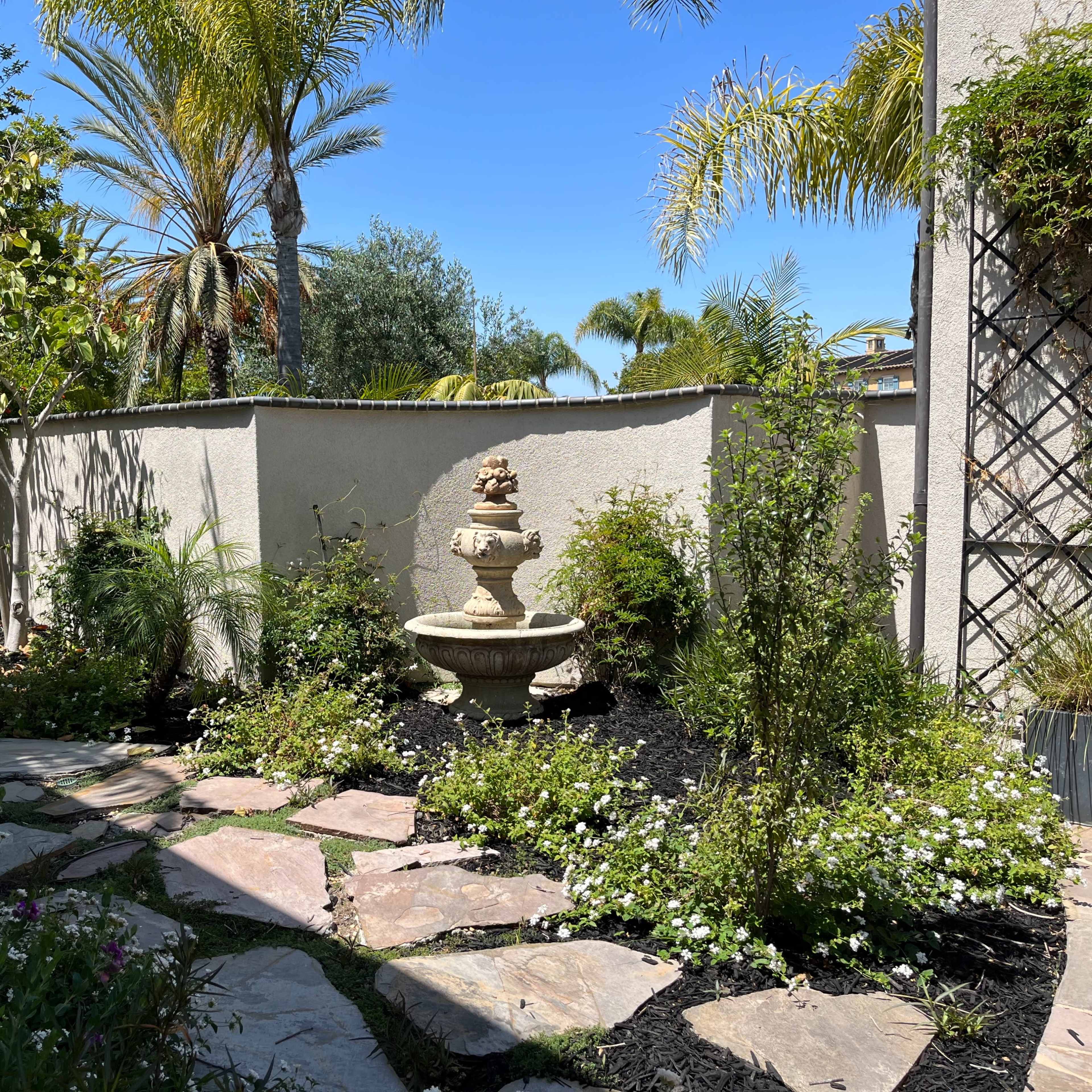 A stone fountain is surrounded by a path of flagstones and lush greenery in a sunlit garden.