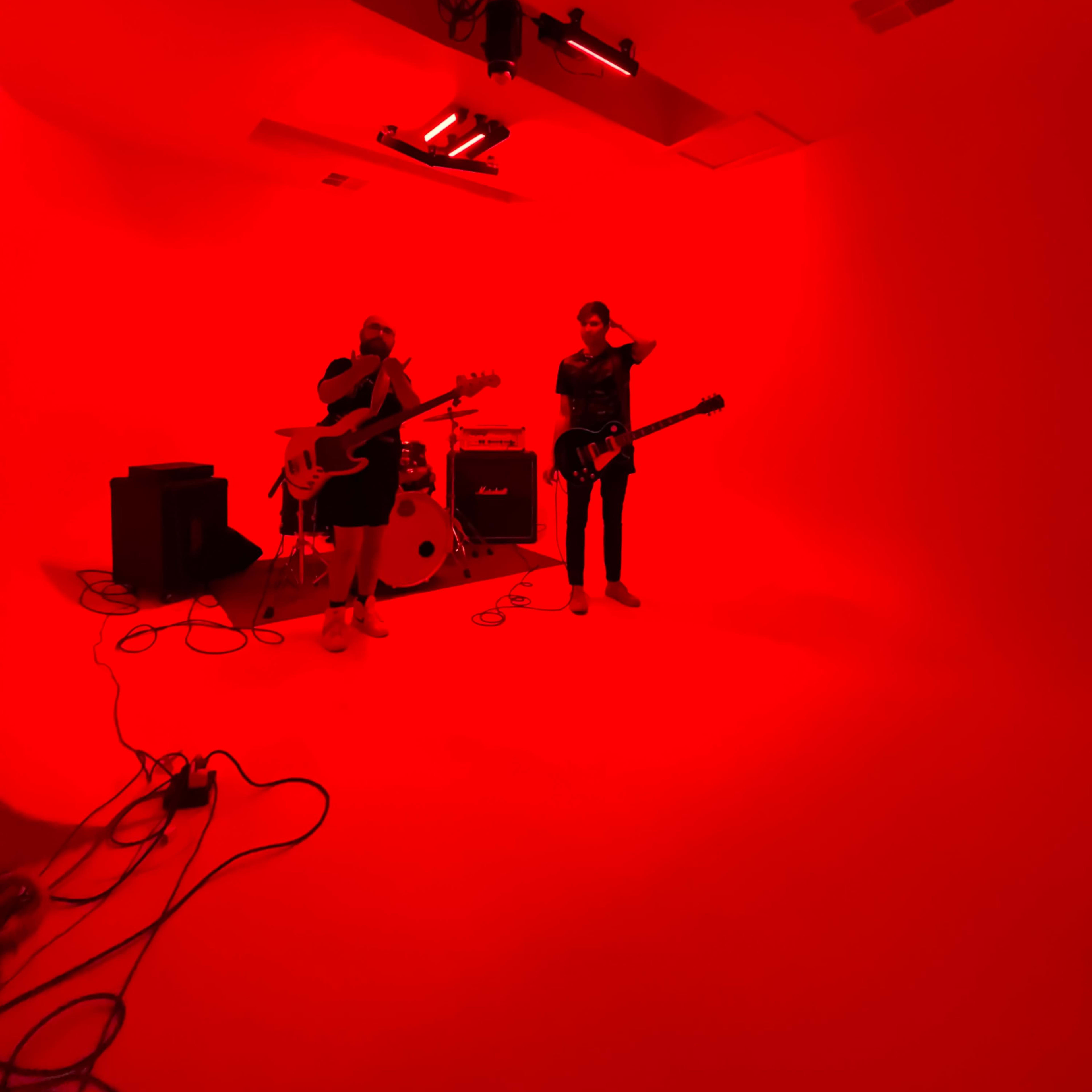 Two musicians are standing in a red-lit studio, one playing a bass guitar and the other holding an electric guitar.