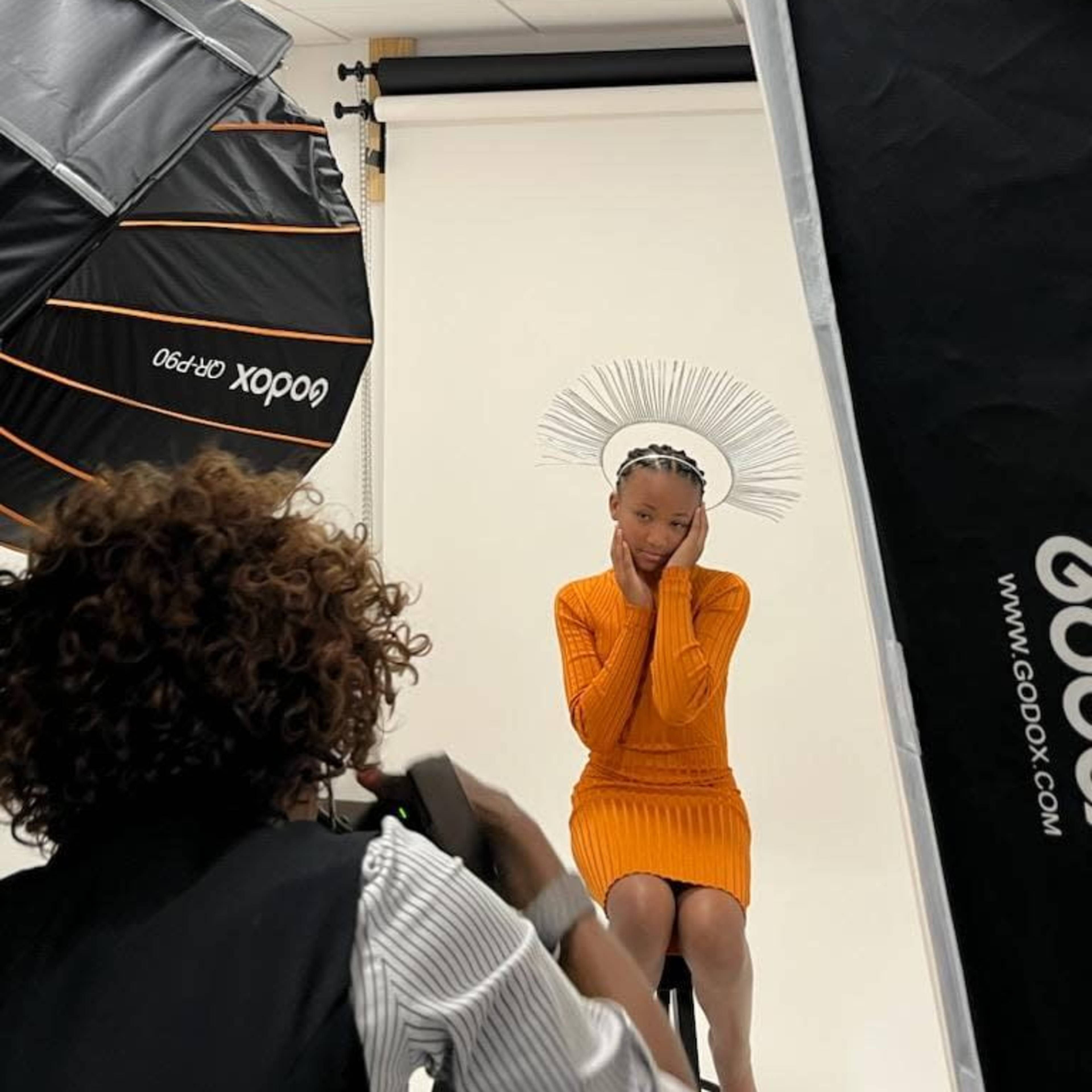A young model in an orange dress poses with her hands on her head in a photography studio, surrounded by large softbox lights.